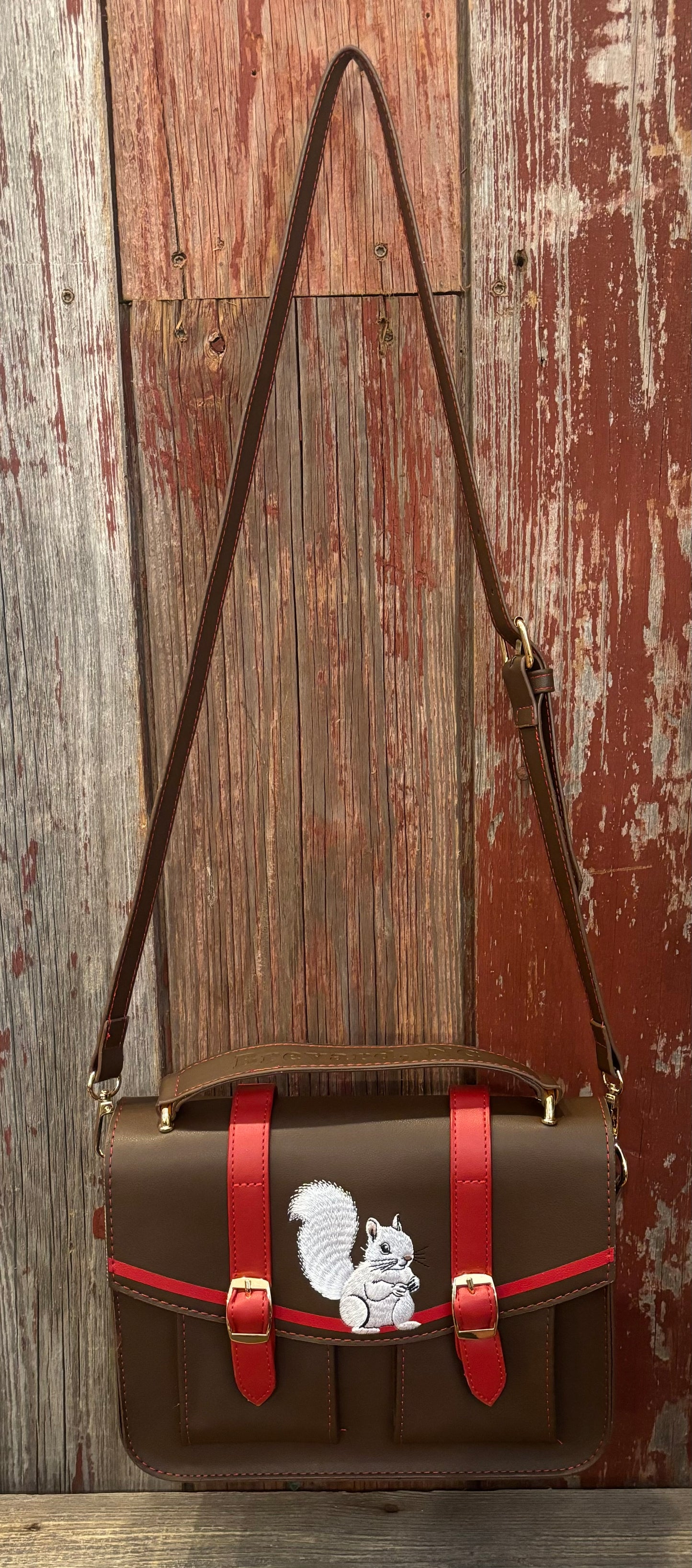 A brown messenger bag with red straps featuring an embroidered white squirrel on the front, with a short handle and long strap. Shown hanging full length..