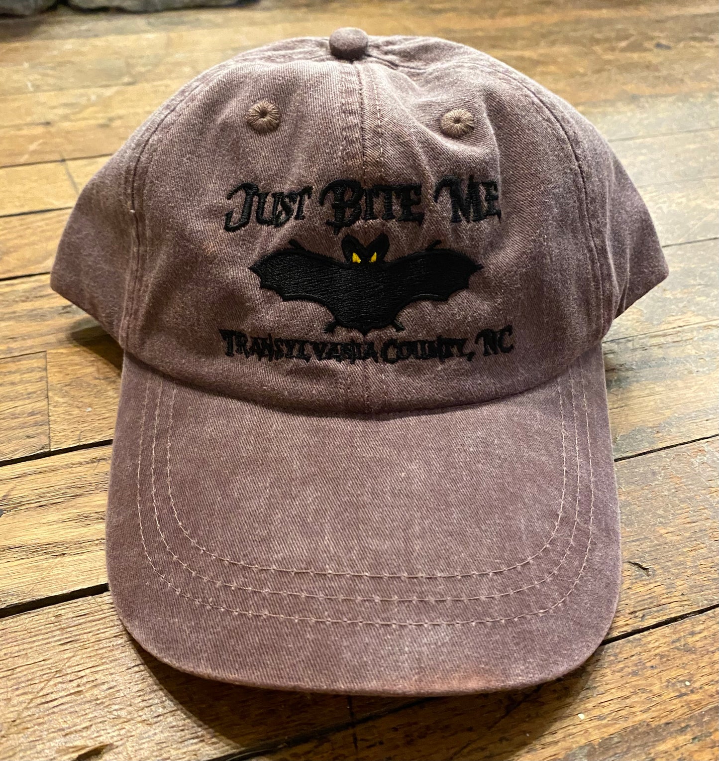 A brown baseball cap with 'Just Bite Me' embroidered on the front and a bat silhouette, indicating a location connection to Transylvania County, NC.