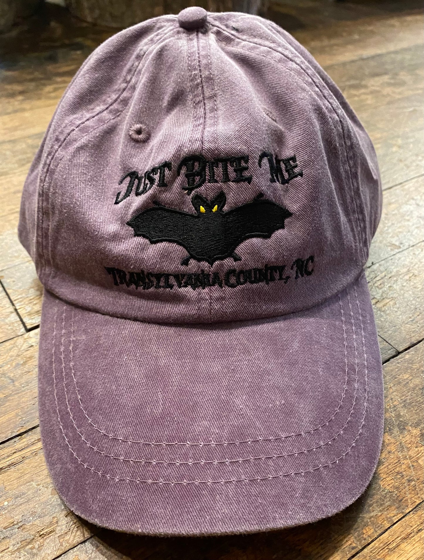 A purple mulberry baseball cap with 'Just Bite Me' embroidered on the front and a bat silhouette, indicating a location connection to Transylvania County, NC.