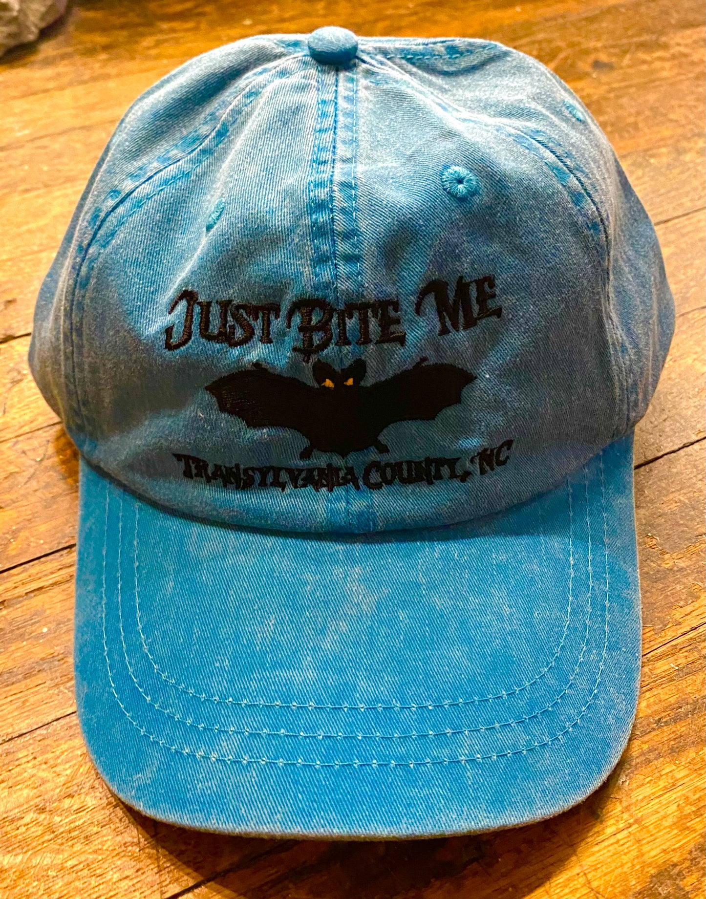 A carribean blue baseball cap with 'Just Bite Me' embroidered on the front and a bat silhouette, indicating a location connection to Transylvania County, NC.