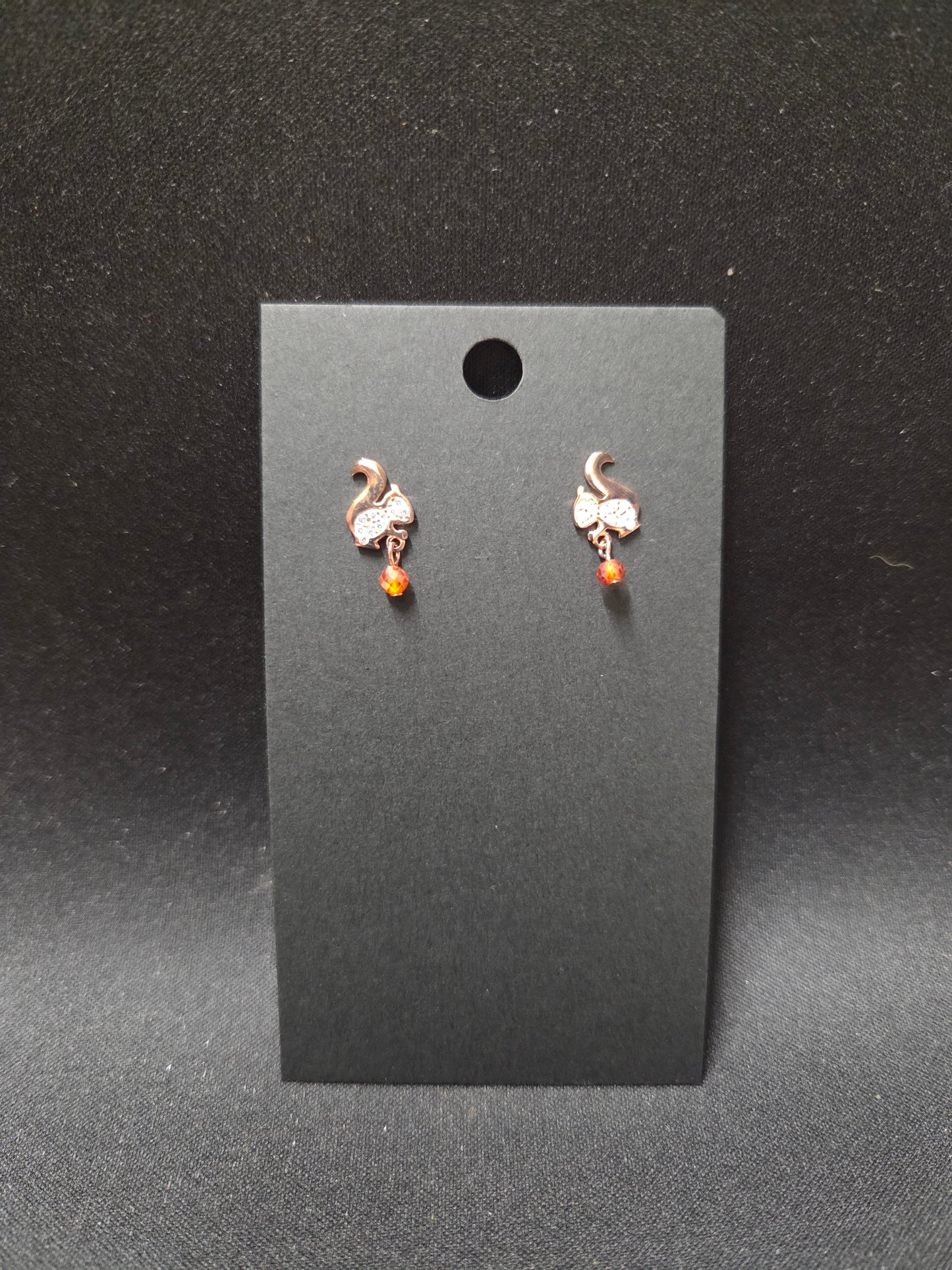 Pair of small stud earrings with a white squirrel and a gem for the nut he's carrying.on a black card against a dark background