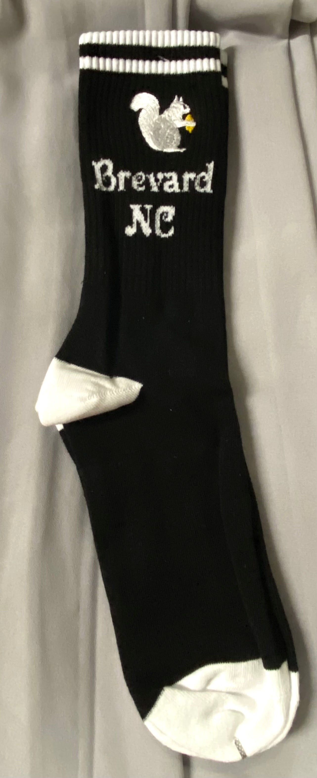 A pair of black crew socks with a white embroidered squirrel design and the text 'Brevard NC' on the cuff.