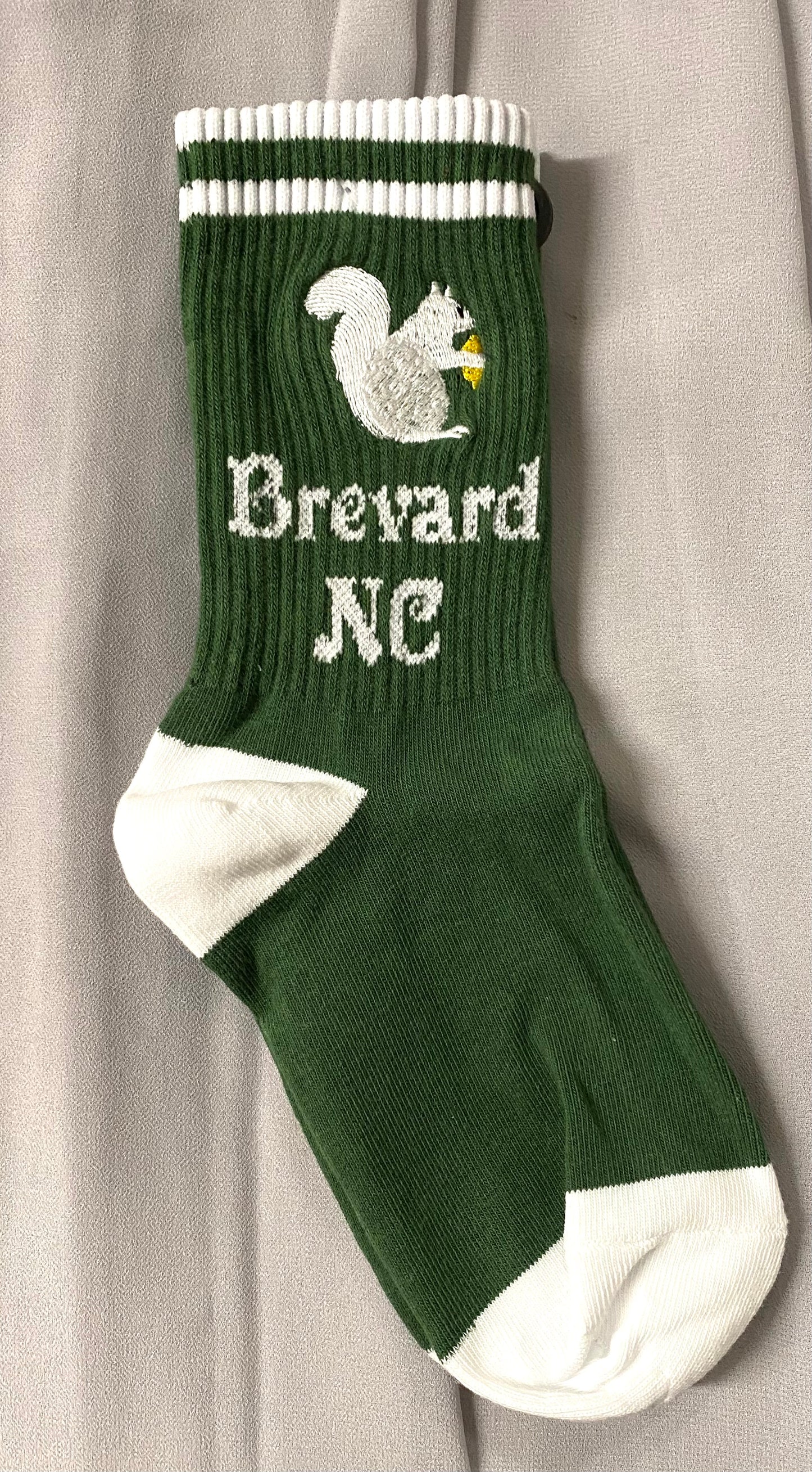 A pair of green crew socks with a white embroidered squirrel and the text 'Brevard NC' in white.