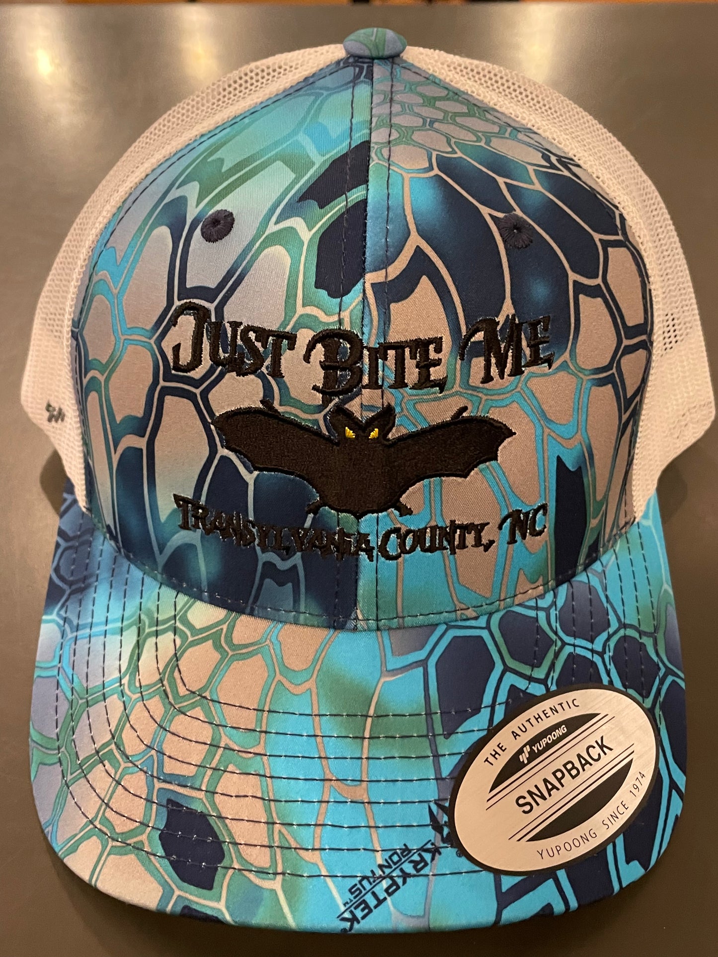 Blue and black patterned cap with 'Just Bite Me' text on a metallic surface