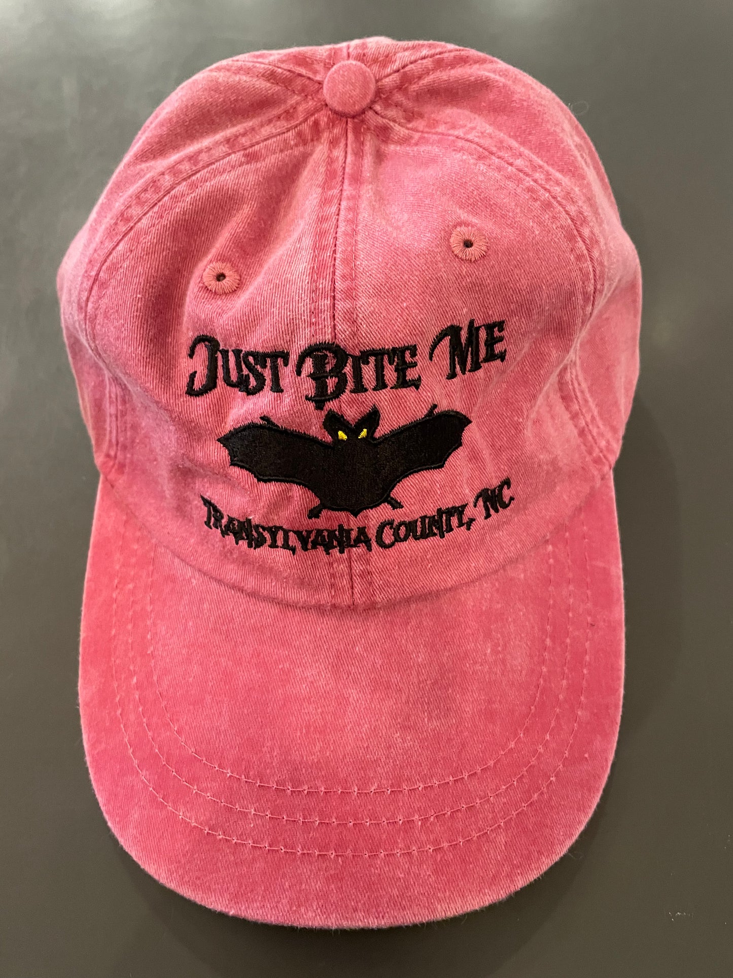 A nautical red baseball cap with 'Just Bite Me' embroidered on the front and a bat silhouette, indicating a location connection to Transylvania County, NC.