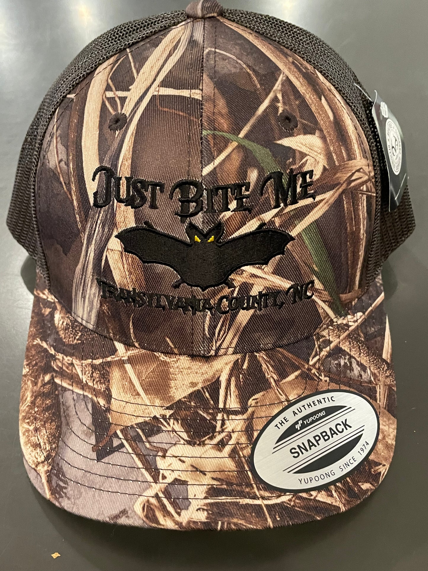 Camouflage cap with 'Just Bite Me' and bat logo on a dark background