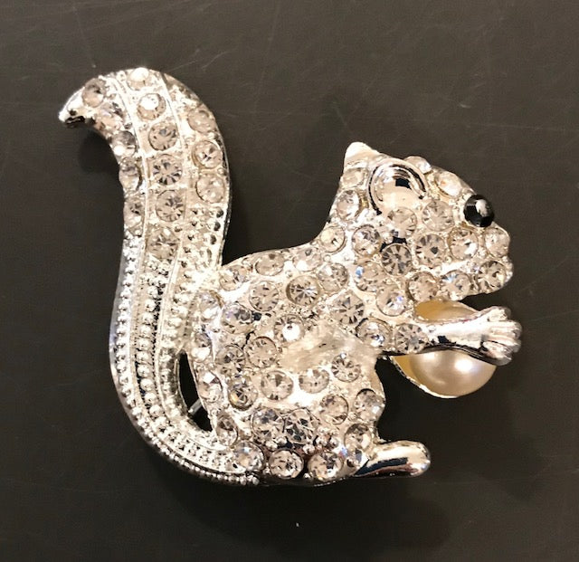 A brooch in the shape of a white squirrel covered with clear rhinestones, with a black rhinestone representing an eye, and a pearl-like substance for an acorn.