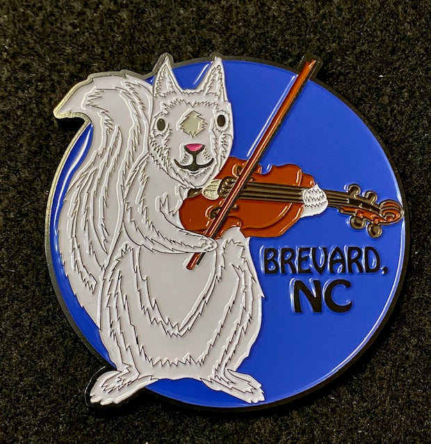 A circular metal magnet featuring an illustration of a white squirrel playing a fiddle with the text 'Brevard, NC' next to it.