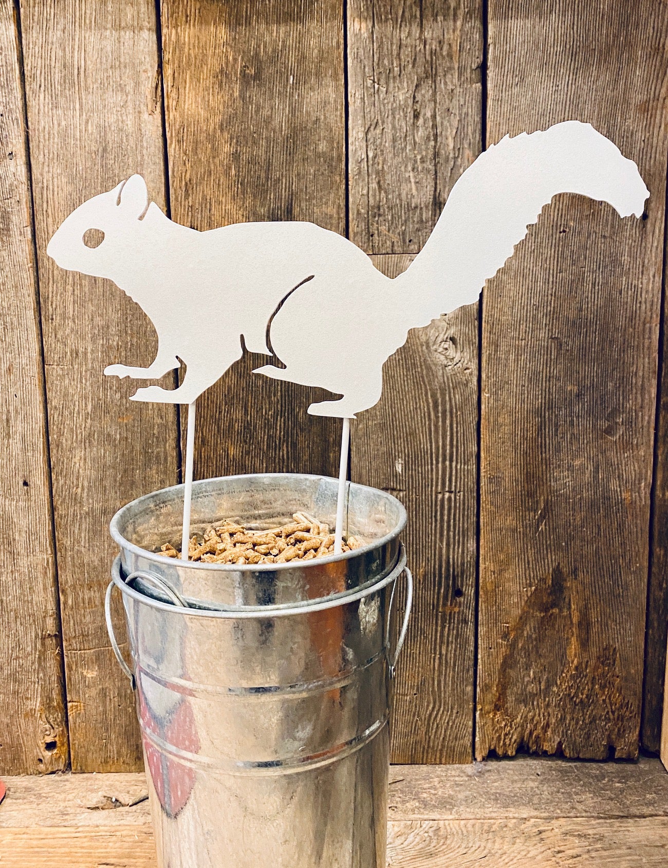 A metal garden stake in the shape of a white squirrel, displayed against a wooden backdrop.
