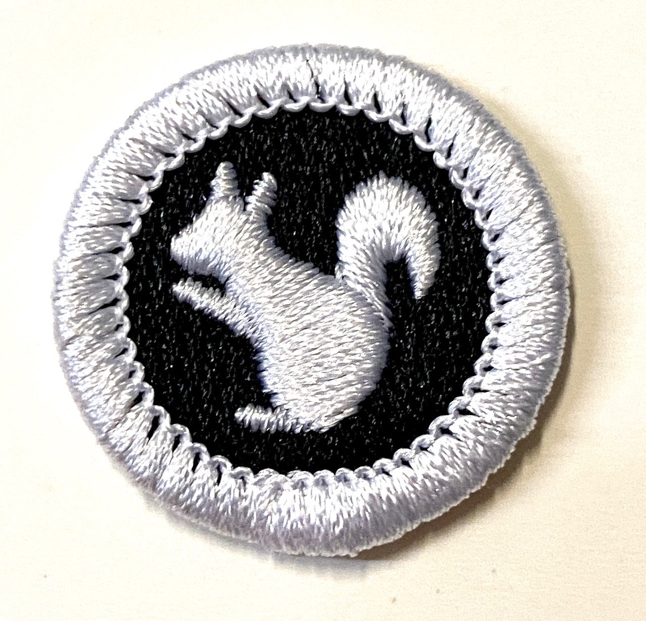 A round embroidered patch with a white squirrel design on a black background with a white trim around the outside.