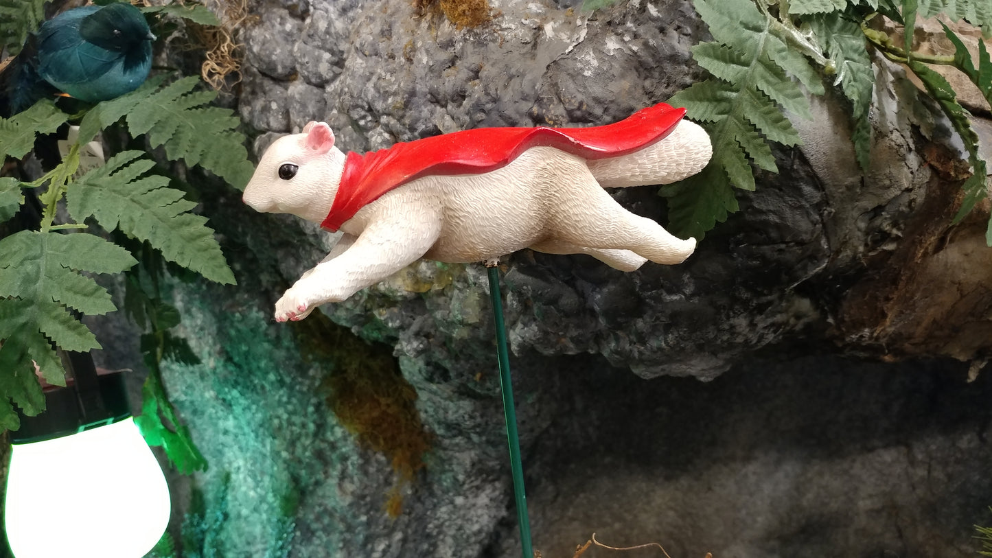 A white squirrel figurine with a red cape attached to a green garden stake.