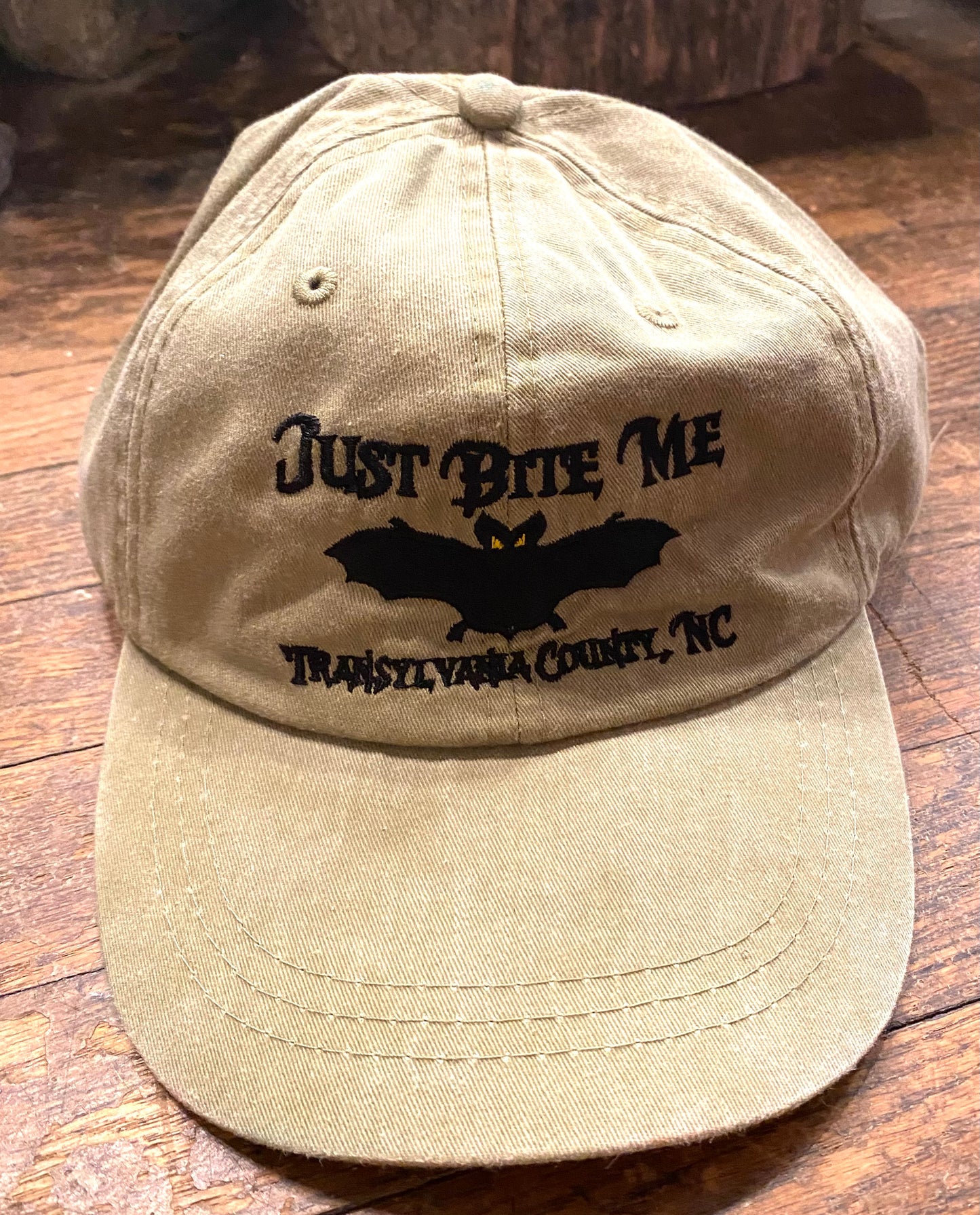 A beige baseball cap with 'Just Bite Me' embroidered on the front and a bat silhouette and Transylvania County, NC.