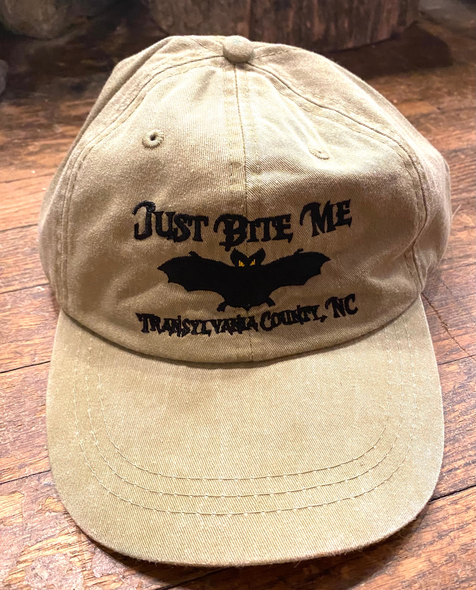 A beige baseball cap with 'Just Bite Me' embroidered on the front and a bat silhouette and Transylvania County, NC.