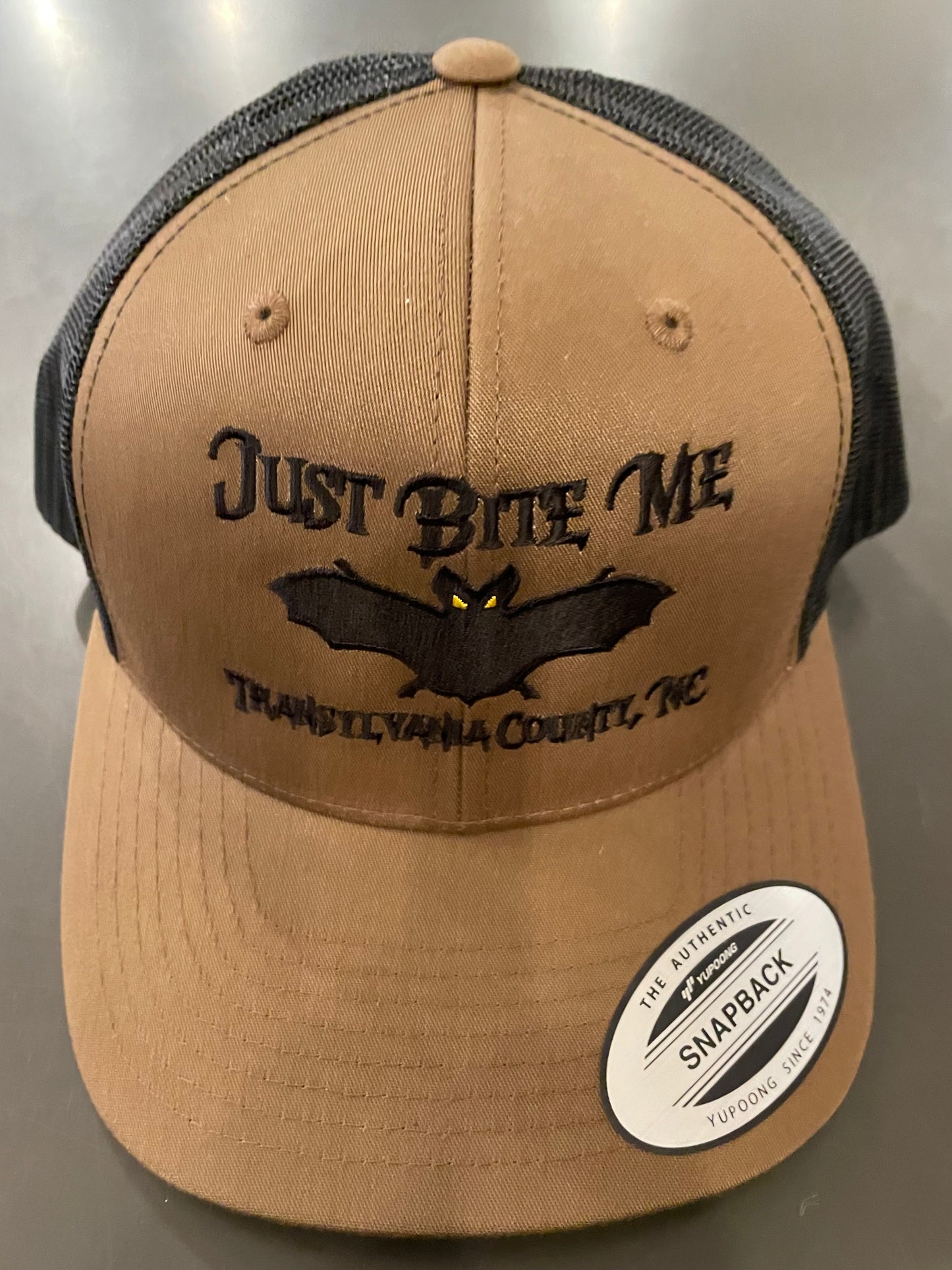 A coyote brown and black baseball cap with 'Just Bite Me' embroidered on the front and a bat silhouette and Transylvania County, NC.