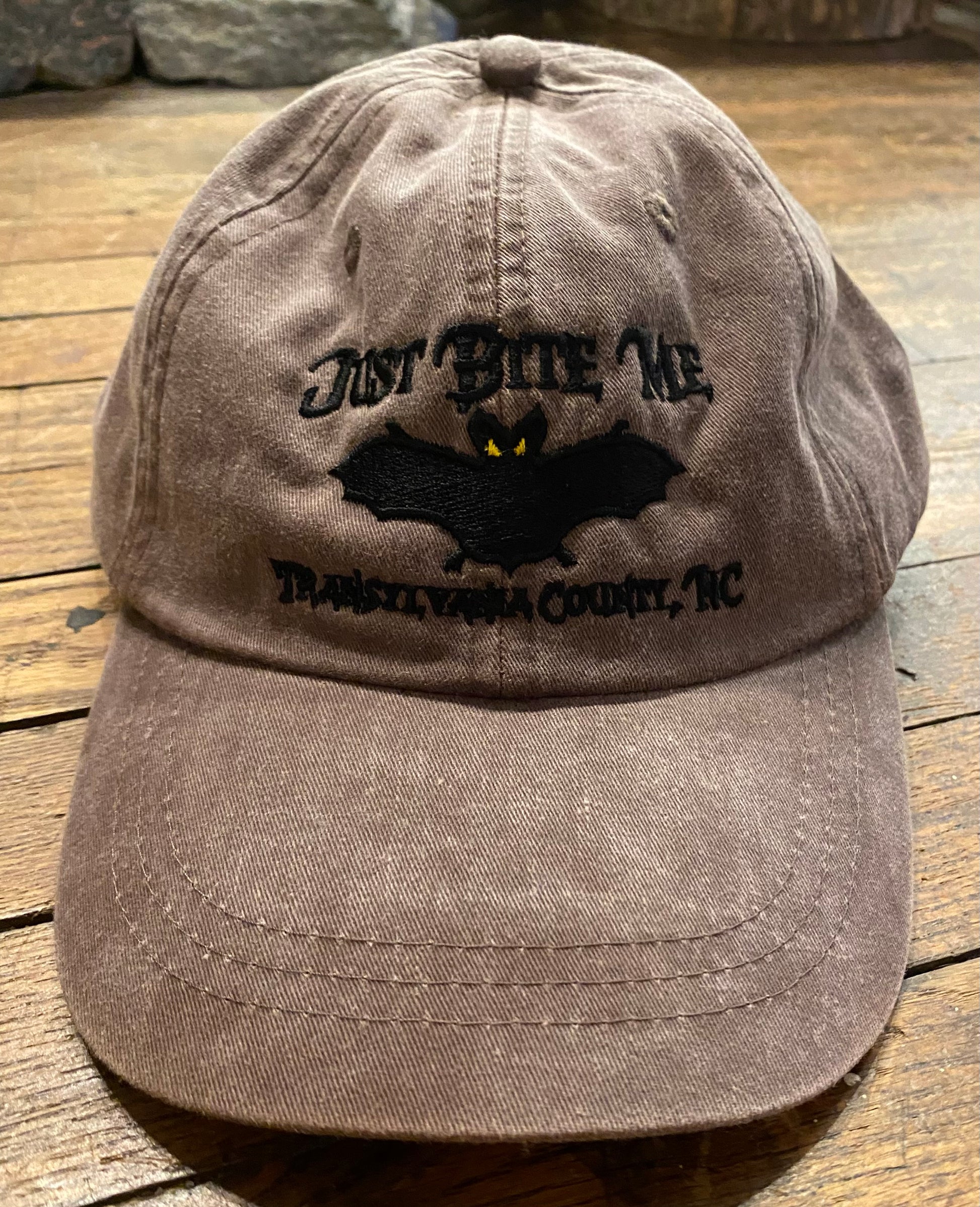 A espresso brown baseball cap with 'Just Bite Me' embroidered on the front and a bat silhouette and Transylvania County, NC.