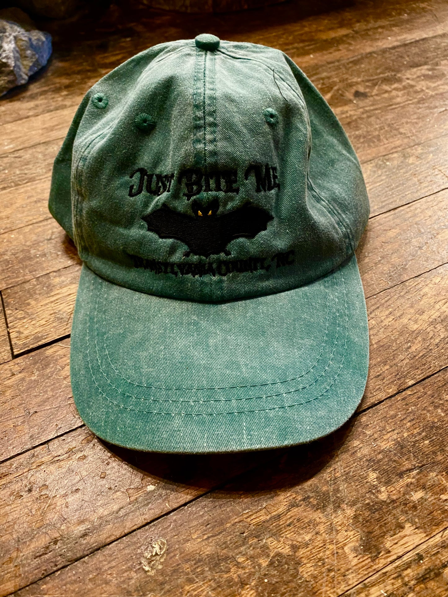 A forest green baseball cap with 'Just Bite Me' embroidered on the front and a bat silhouette and Transylvania County, NC.