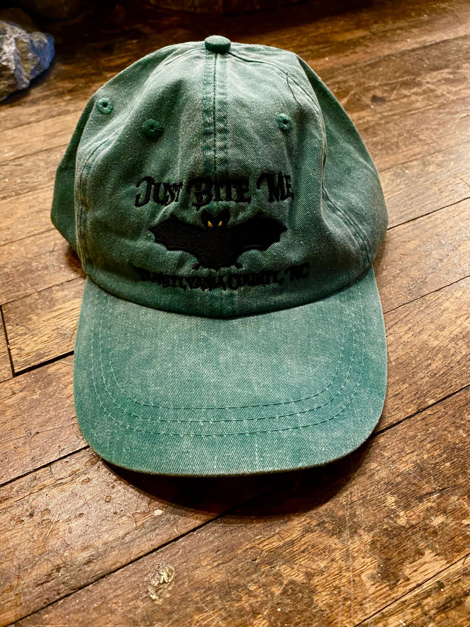 A forest green baseball cap with 'Just Bite Me' embroidered on the front and a bat silhouette and Transylvania County, NC.