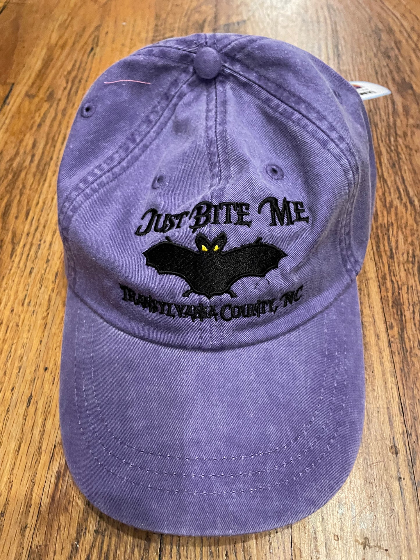 A grape purple baseball cap with 'Just Bite Me' embroidered on the front and a bat silhouette and Transylvania County, NC.