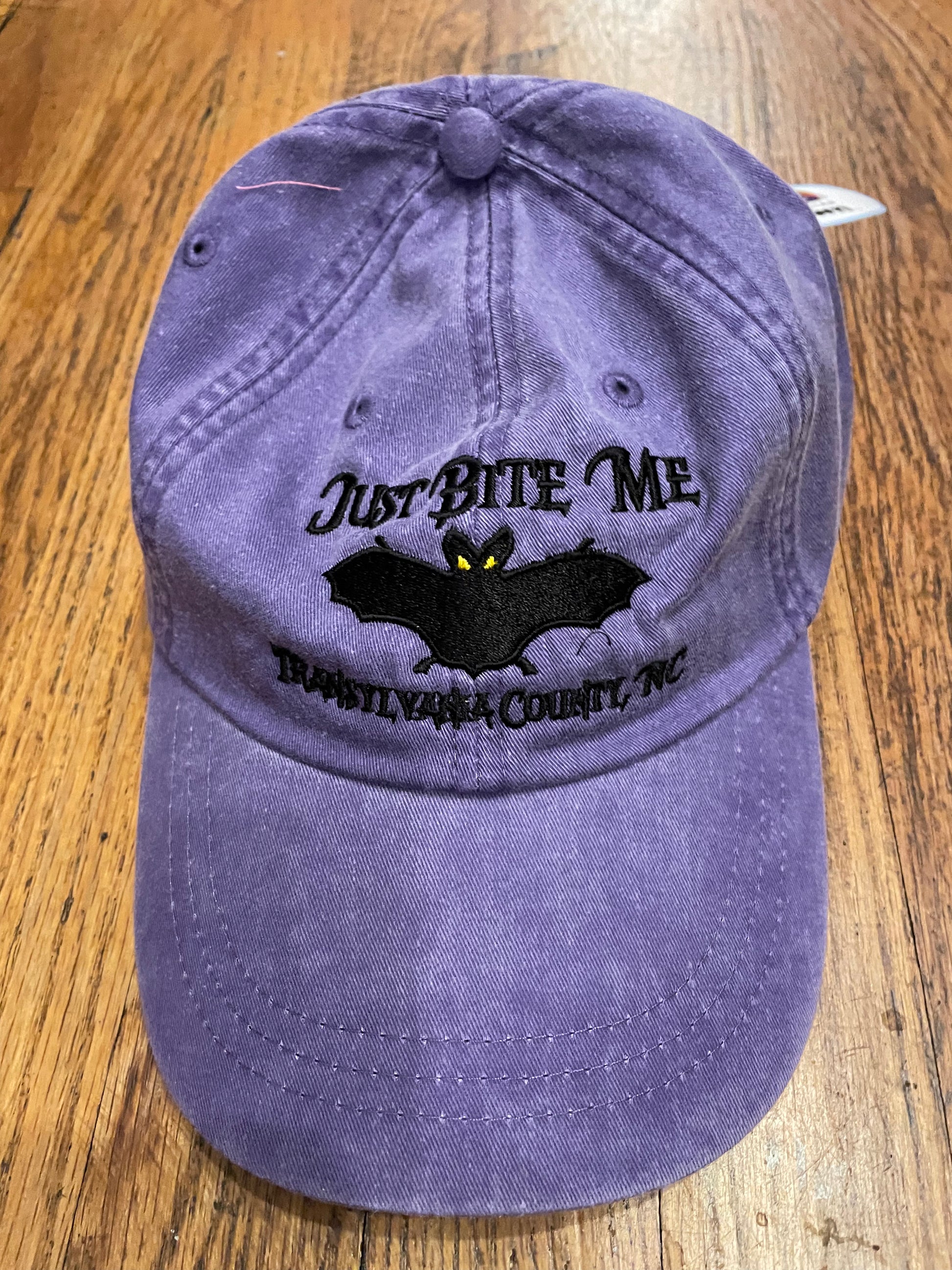 A grape purple baseball cap with 'Just Bite Me' embroidered on the front and a bat silhouette and Transylvania County, NC.