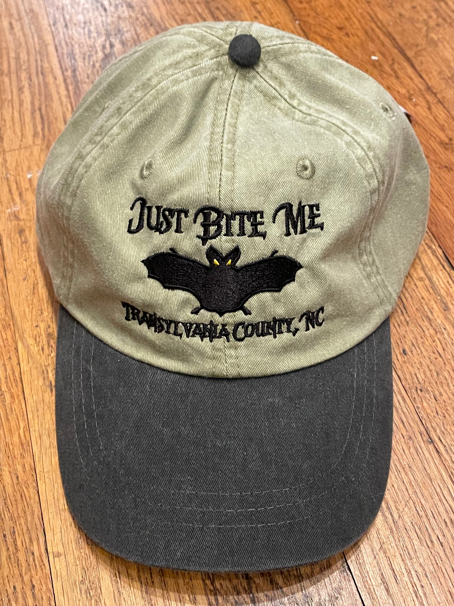 A khaki and black baseball cap with 'Just Bite Me' embroidered on the front and a bat silhouette and Transylvania County, NC.