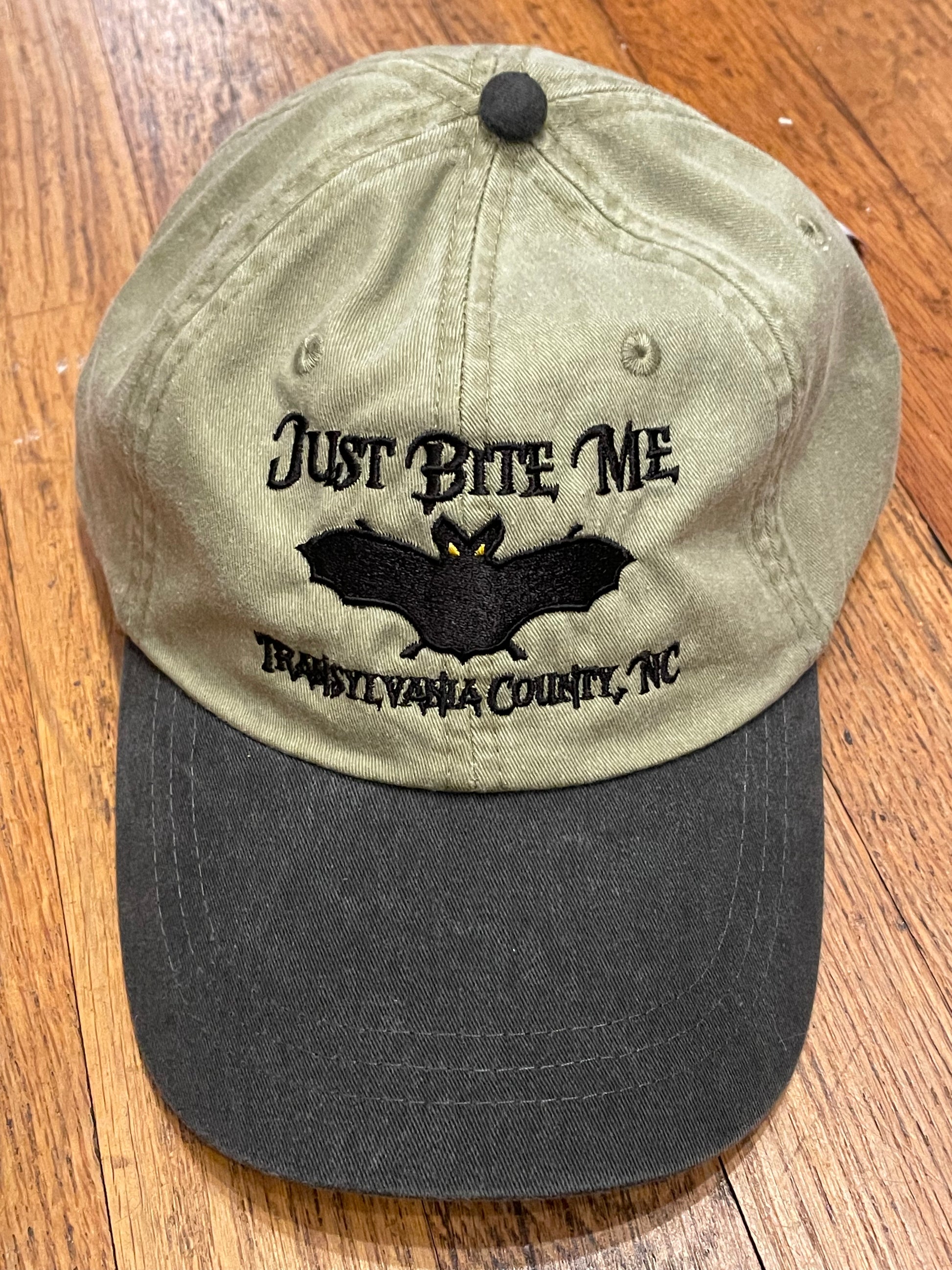 A khaki and black baseball cap with 'Just Bite Me' embroidered on the front and a bat silhouette and Transylvania County, NC.