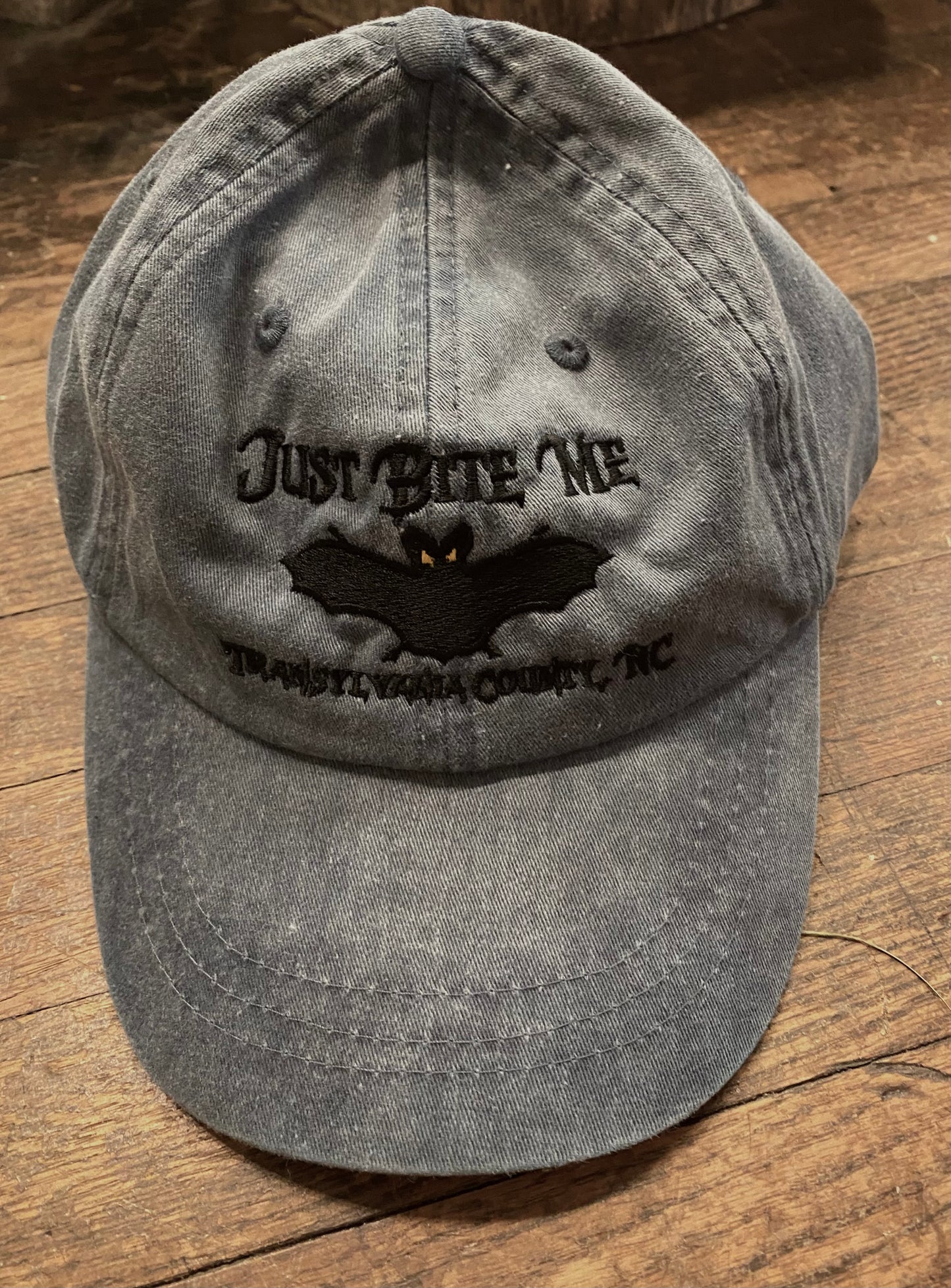 A light grey baseball cap with 'Just Bite Me' embroidered on the front and a bat silhouette and Transylvania County, NC.