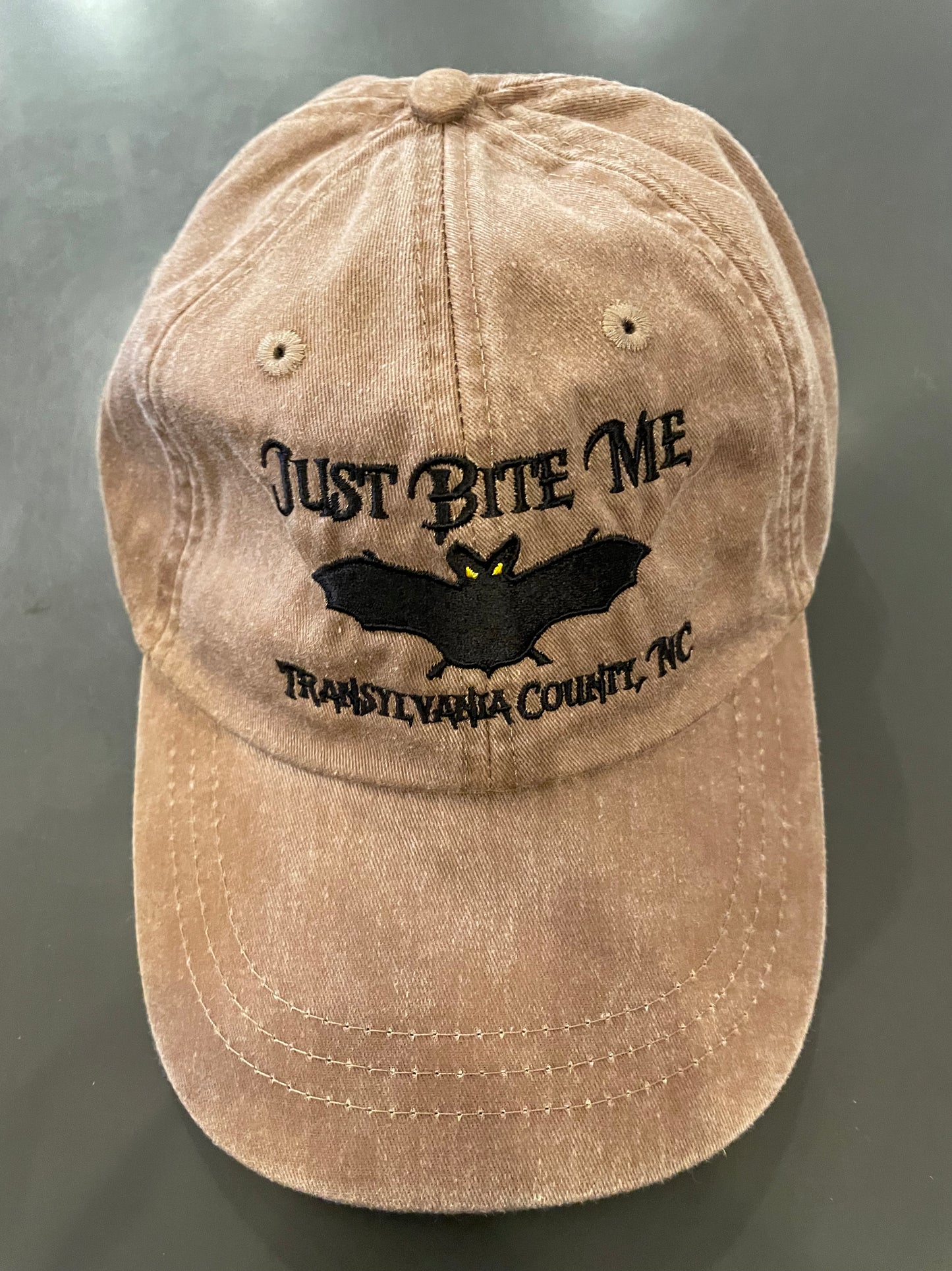 A Mississippi mud brown baseball cap with 'Just Bite Me' embroidered on the front and a bat silhouette and  Transylvania County, NC.