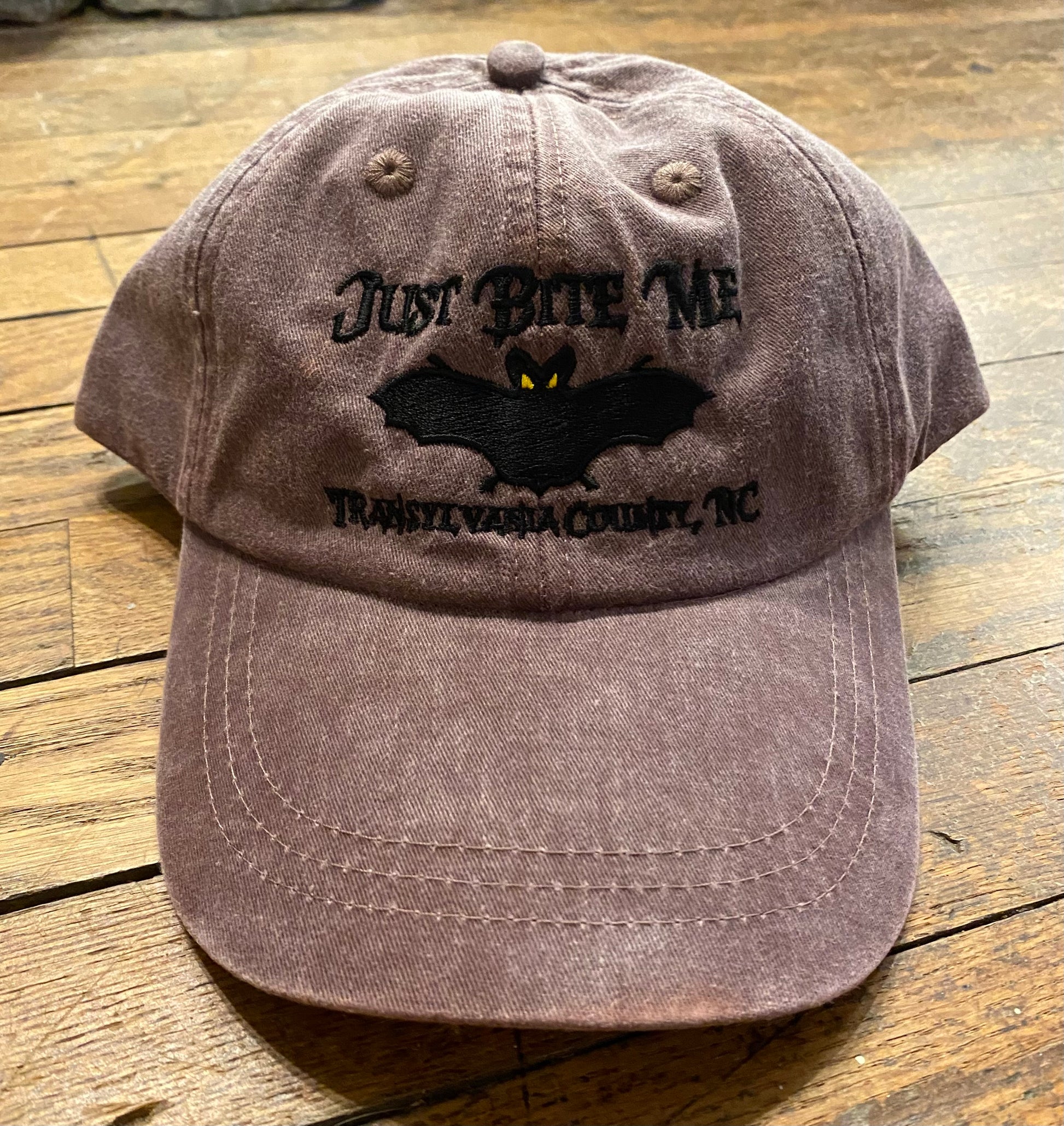 A mulberry baseball cap with 'Just Bite Me' embroidered on the front and a bat silhouette and Transylvania County, NC.