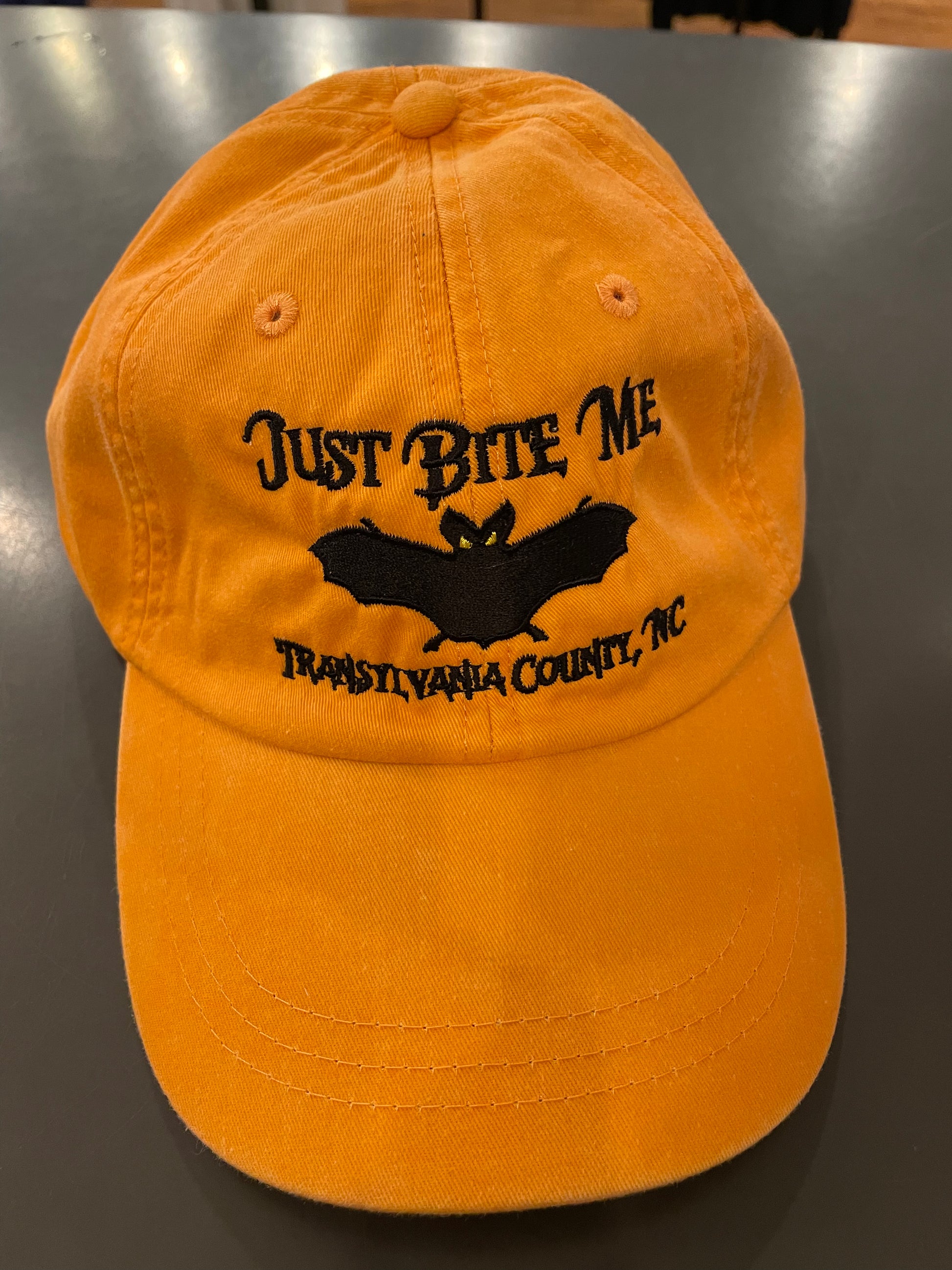 A tangerine orange baseball cap with 'Just Bite Me' embroidered on the front and a bat silhouette and  Transylvania County, NC.