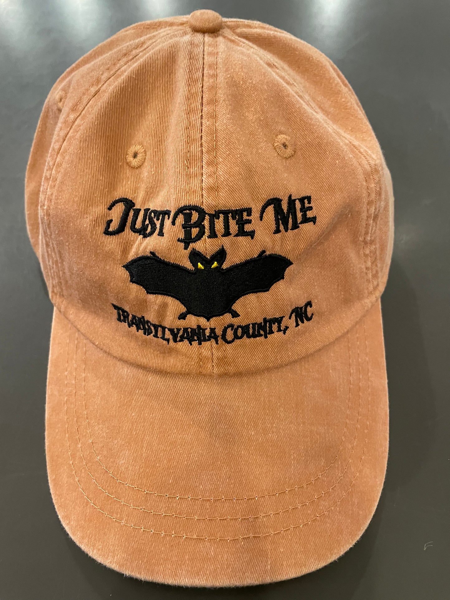 A terra cotta colored baseball cap with 'Just Bite Me' embroidered on the front and a bat silhouette and Transylvania County, NC.