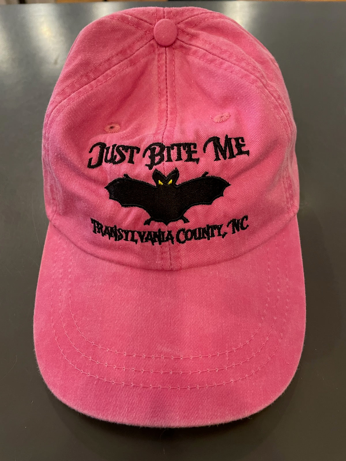 A pink baseball cap with 'Just Bite Me' embroidered on the front and a bat silhouette and Transylvania County, NC.