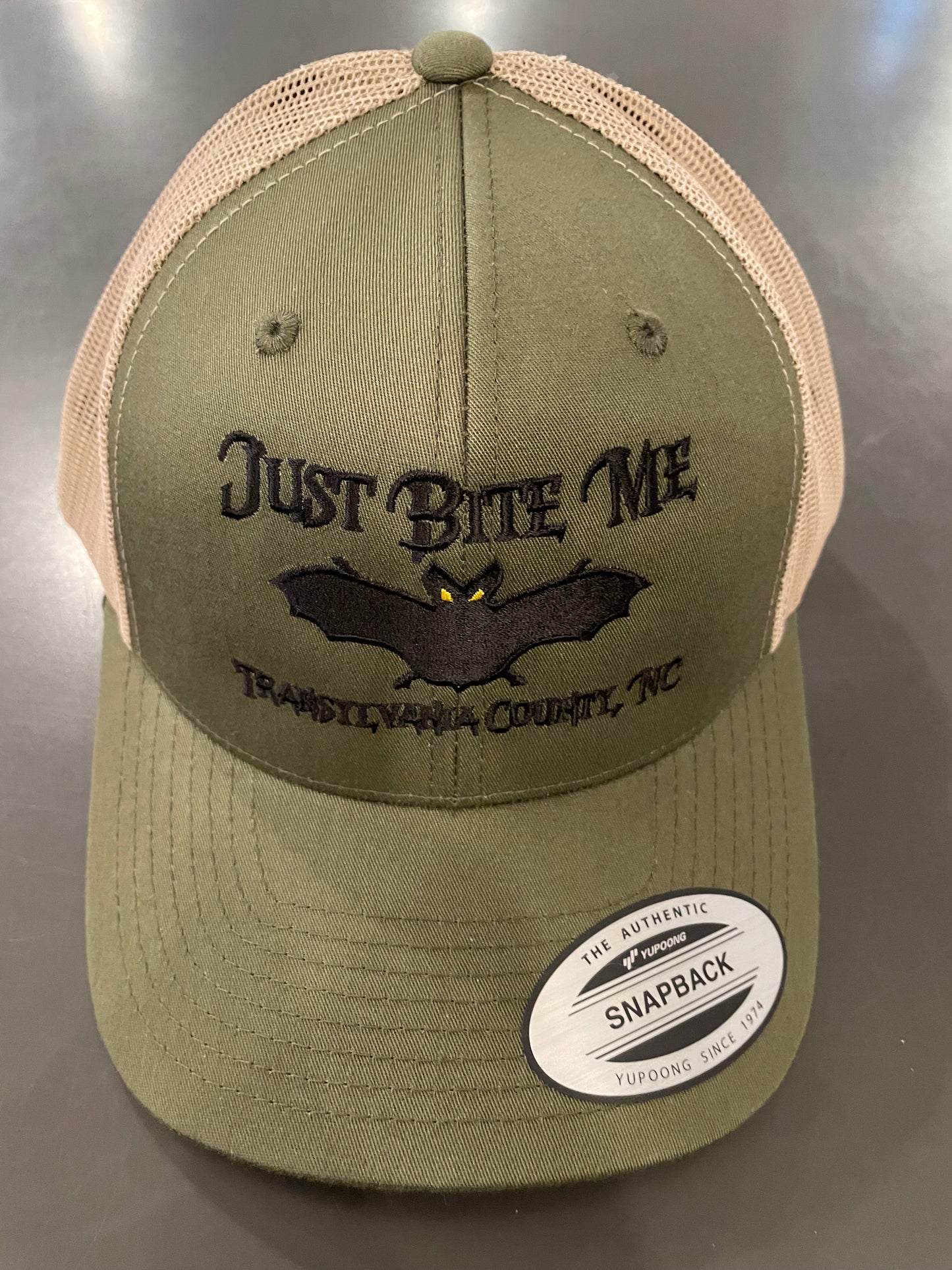 A moss green and khaki  baseball cap with 'Just Bite Me' embroidered on the front and a bat silhouette and Transylvania County, NC.