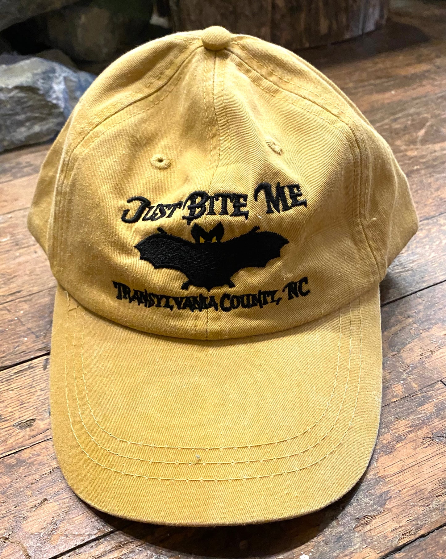 A mustard yellow baseball cap with 'Just Bite Me' embroidered on the front and a bat silhouette and Transylvania County, NC.