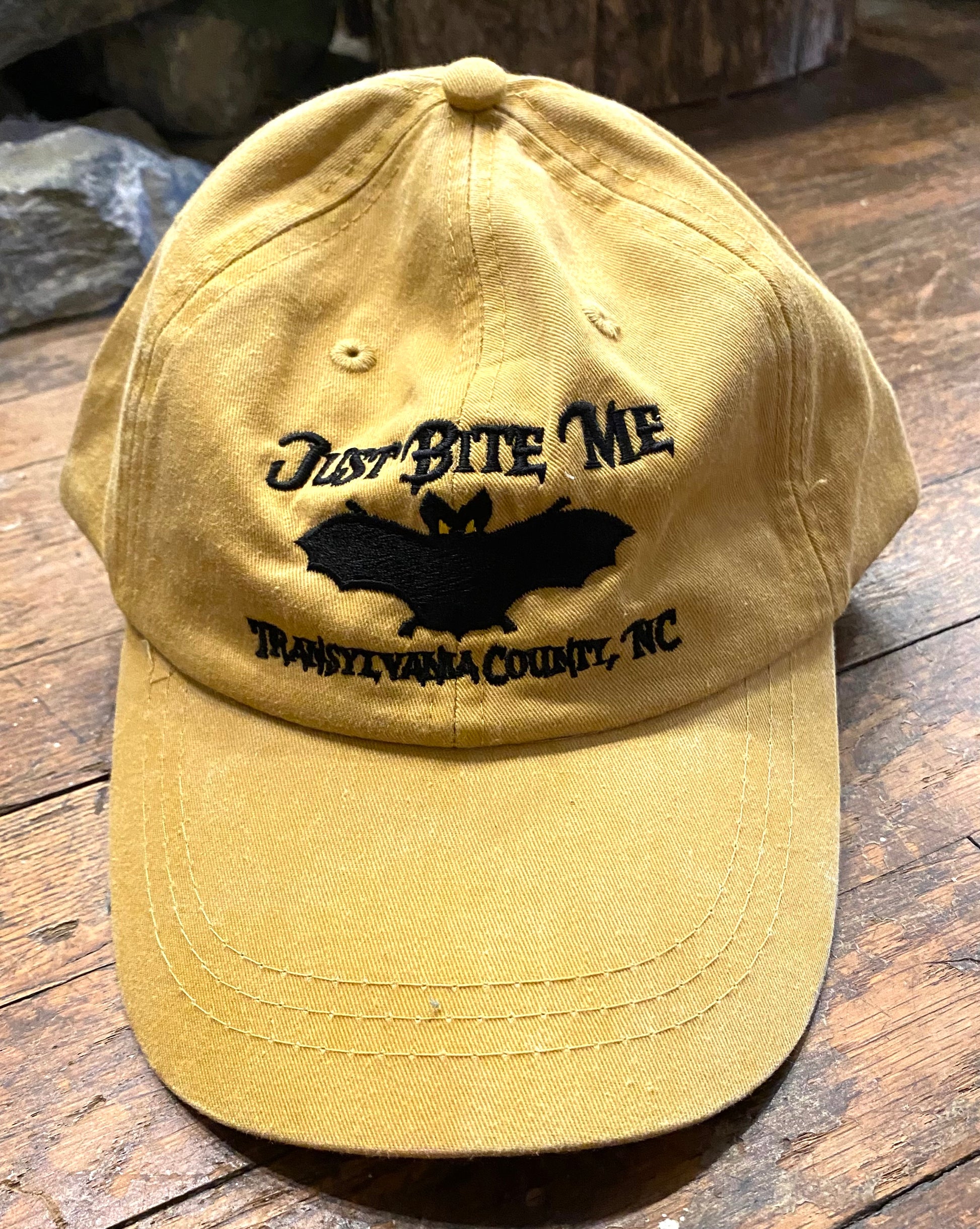 A mustard yellow baseball cap with 'Just Bite Me' embroidered on the front and a bat silhouette and Transylvania County, NC.