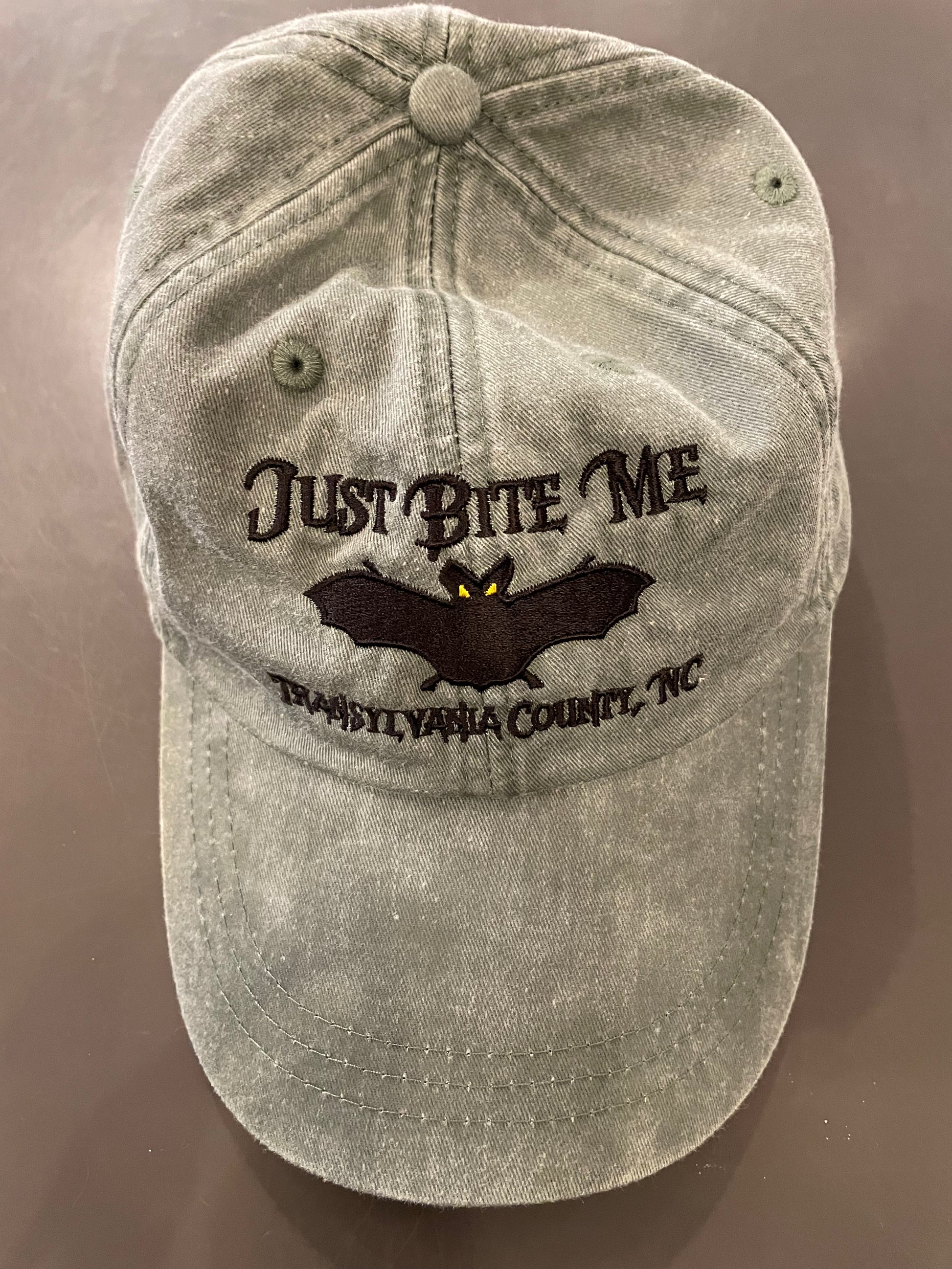 A spruce green baseball cap with 'Just Bite Me' embroidered on the front and a bat silhouette and Transylvania County, NC.