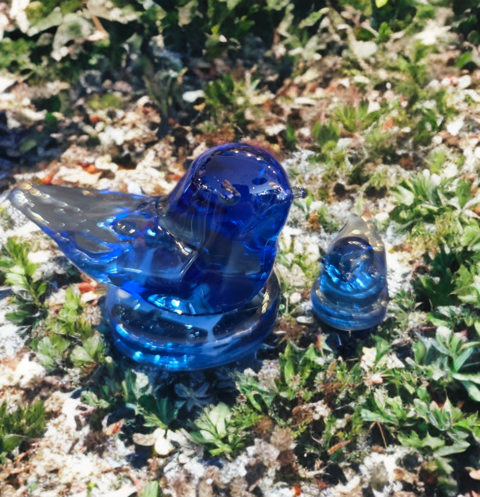 Two blue glass bird figurines on a natural ground with greenery.