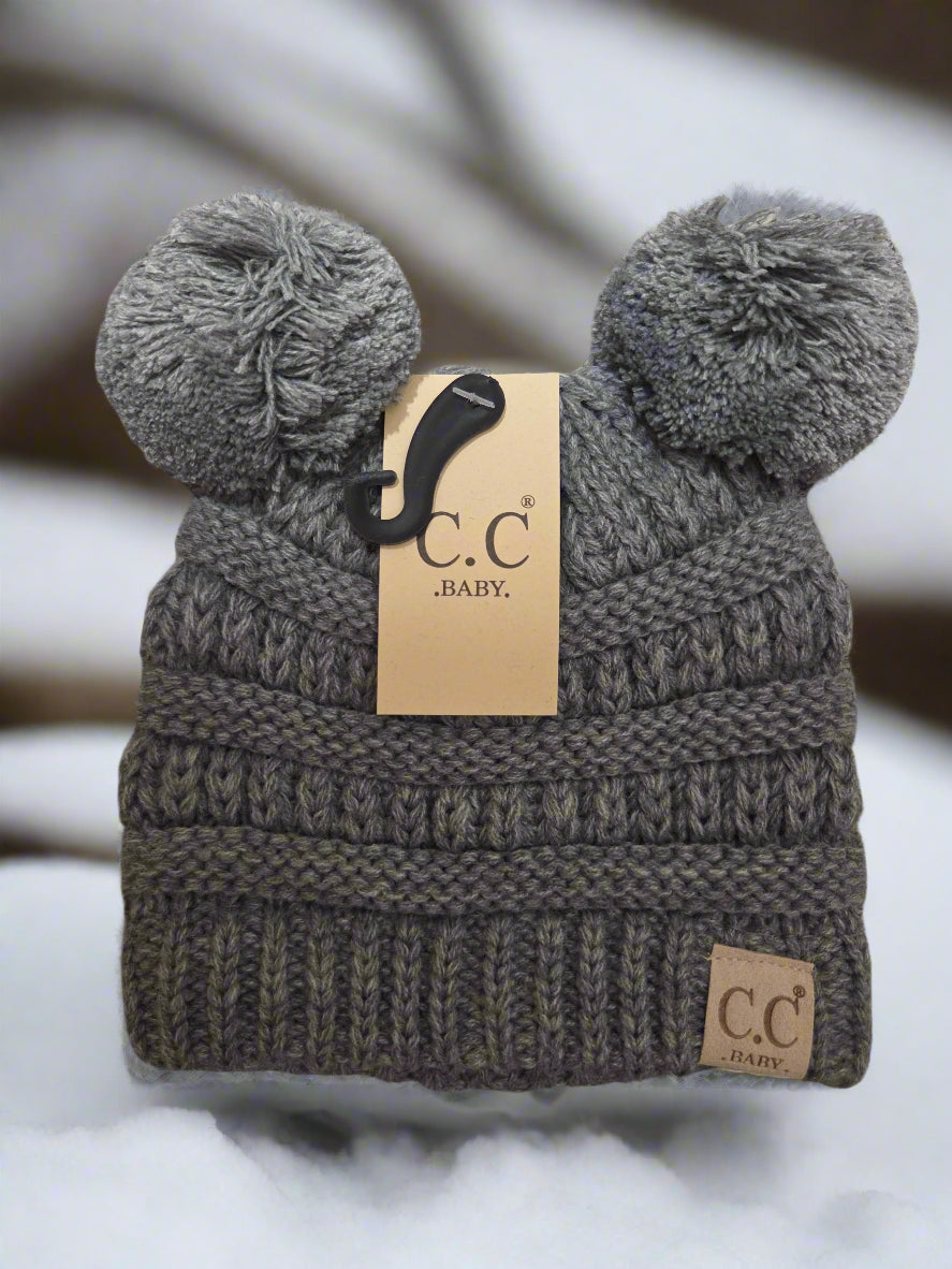 Gray knit baby hat with pom-poms and a matching blanket on a textured surface, featuring a brand tag.