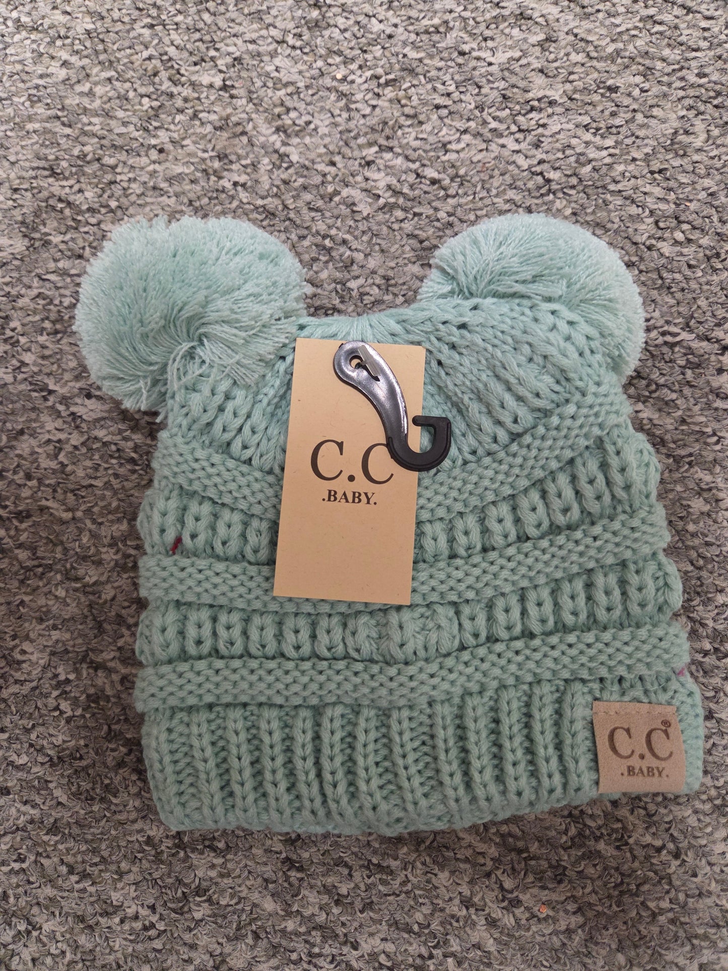 Light green knitted baby hat with pom-poms and a visible brand tag on a gray carpet.