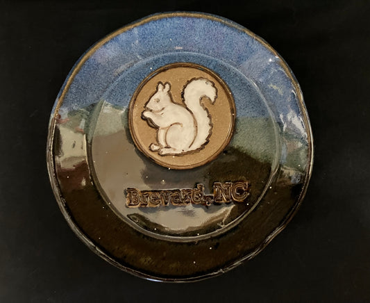 A handmade pottery plate featuring a blue speckled glaze, a brown central medallion with a white squirrel silhouette, and the embossed text 'Brevard, NC' on a dark background.