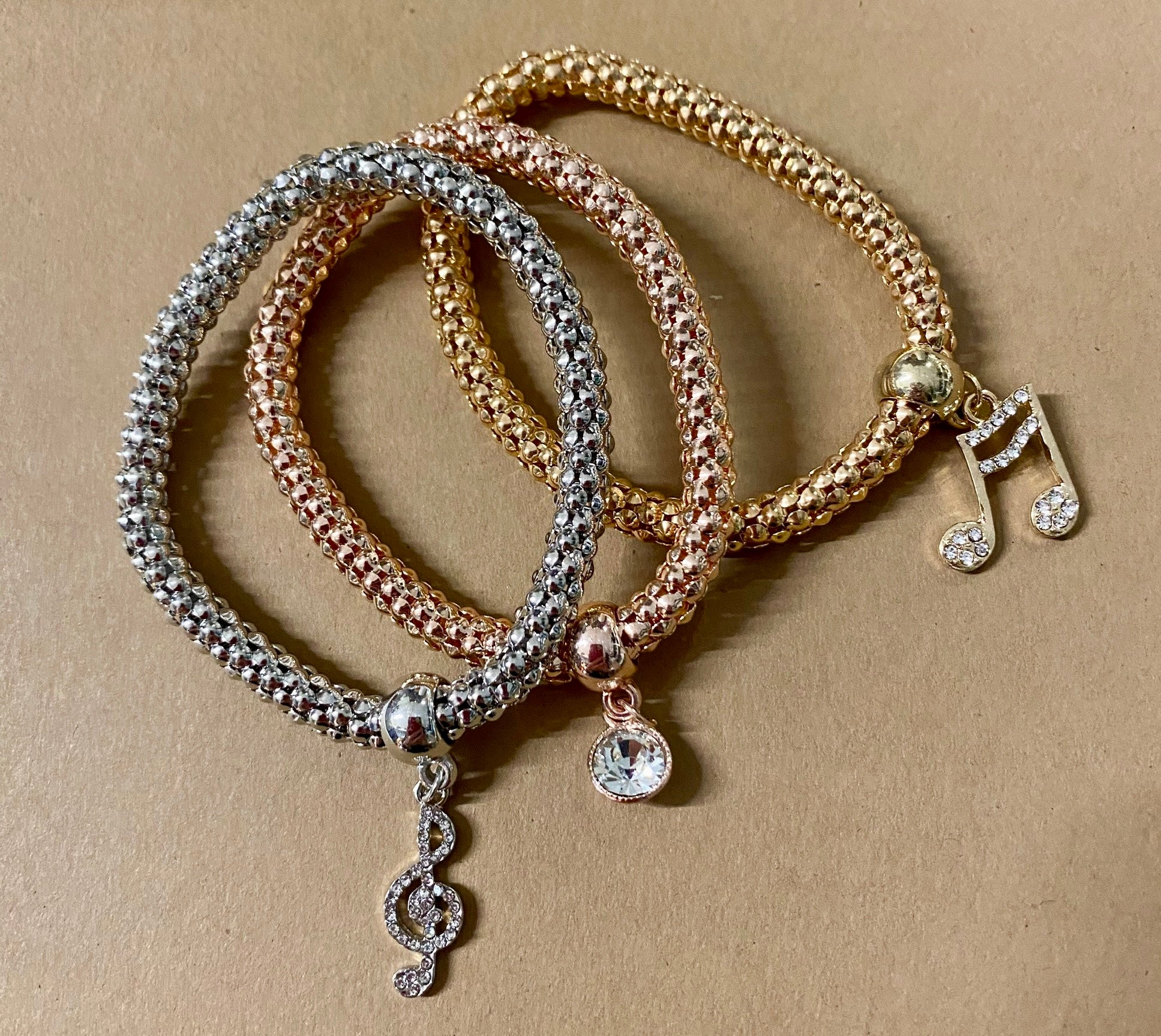 Three bracelets laid out on a surface, featuring gold, silver, and rose gold colors with rhinestone musical charms on each.