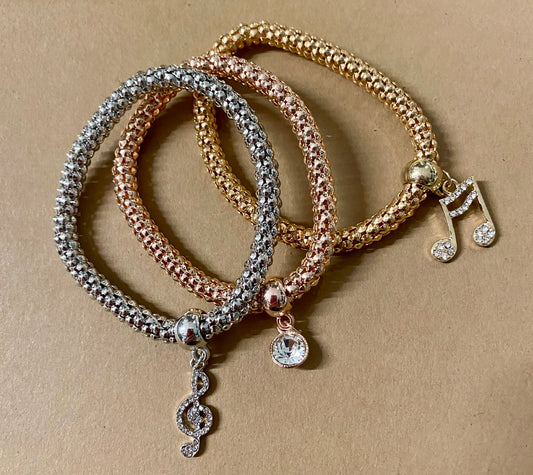 Three bracelets laid out on a surface, featuring gold, silver, and rose gold colors with rhinestone musical charms on each.