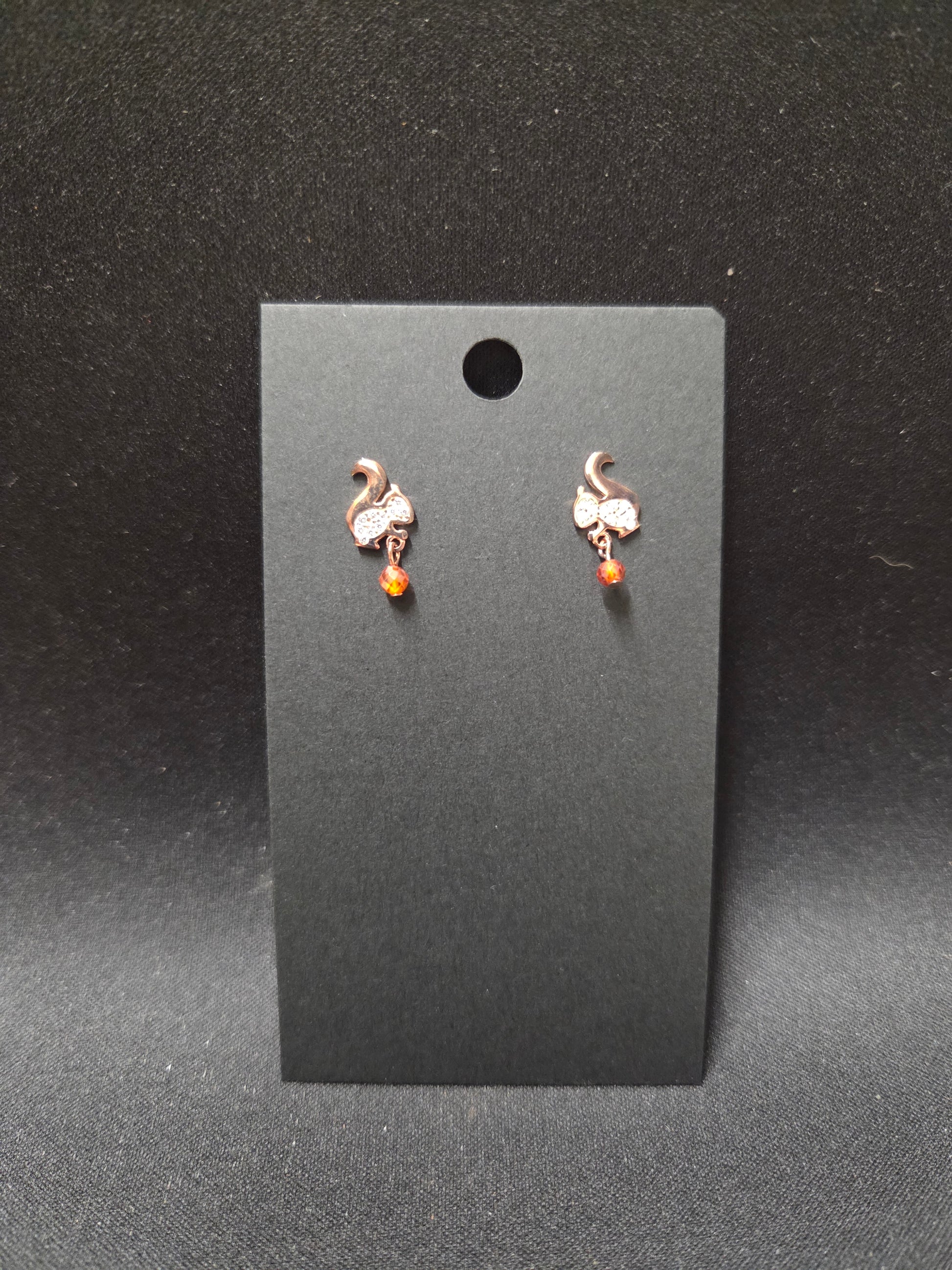 Pair of small stud earrings with a white squirrel and a gem for the nut he's carrying.on a black card against a dark background