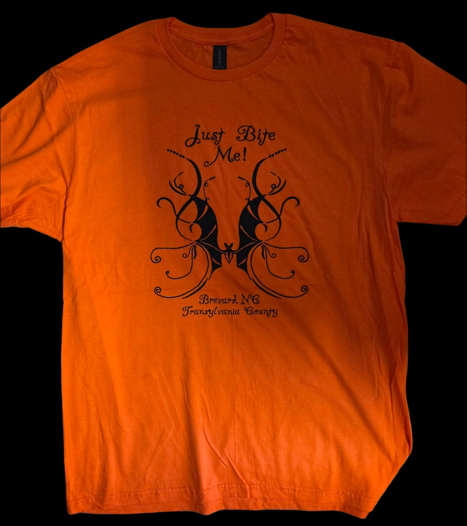 An orange short-sleeve crew neck t-shirt with a black graphic print featuring a swirly bat and the text 'Just Bite Me!', 'Brevard, NC', and 'Transylvania County' on a solid orange background.