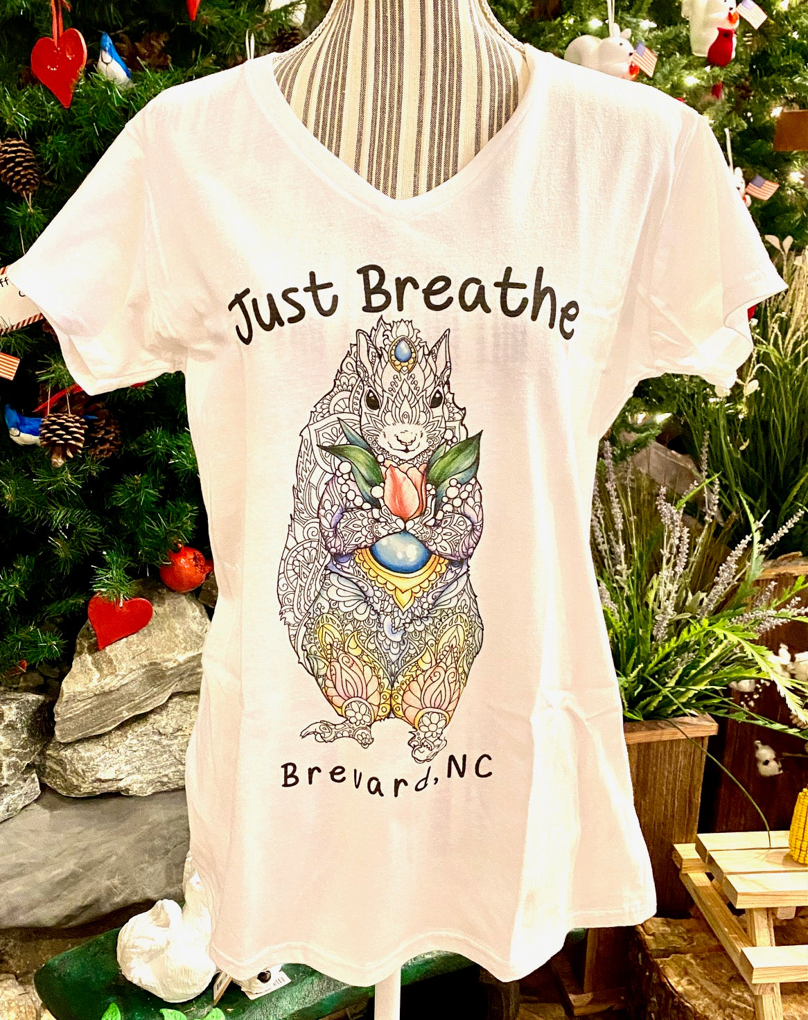 A white, short-sleeved V-neck t-shirt with the text 'Just Breathe' and an illustration of a squirrel, accompanied by the地点 Brevard, NC.