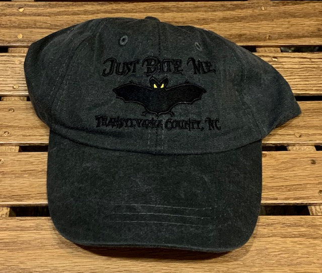 A black baseball cap with 'Just Bite Me' embroidered on the front and a bat silhouette and Transylvania County, NC.