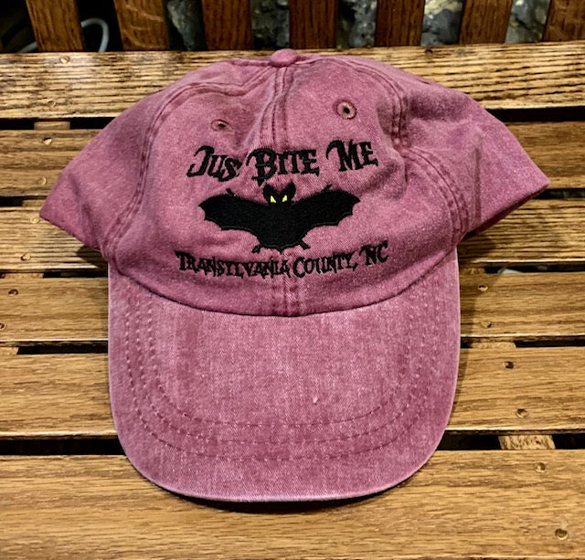 A burgundy baseball cap with 'Just Bite Me' embroidered on the front and a bat silhouette and Transylvania County, NC.