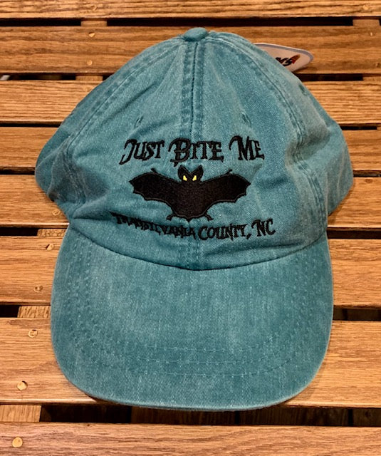 A Caribbean blue green baseball cap with 'Just Bite Me' embroidered on the front and a bat silhouette and Transylvania County, NC.