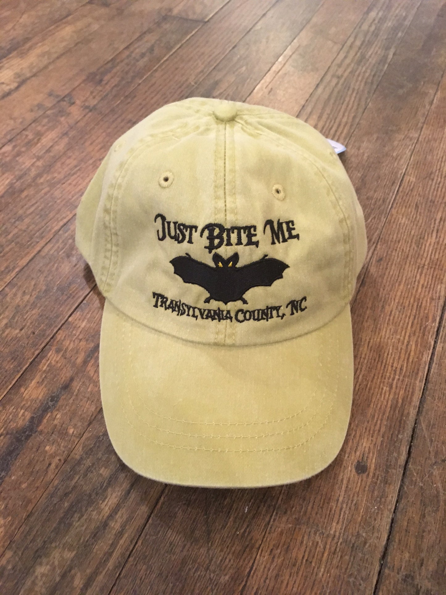 A chamois yellow baseball cap with 'Just Bite Me' embroidered on the front and a bat silhouette and Transylvania County, NC.