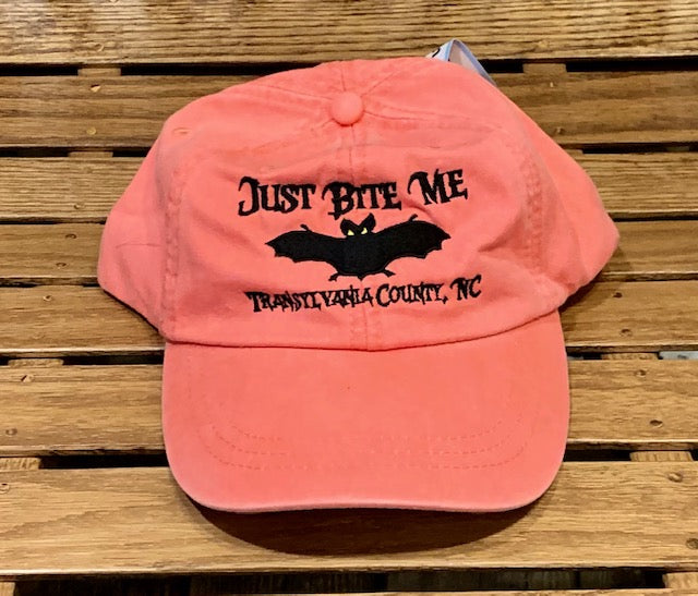 A coral pink baseball cap with 'Just Bite Me' embroidered on the front and a bat silhouette and Transylvania County, NC.