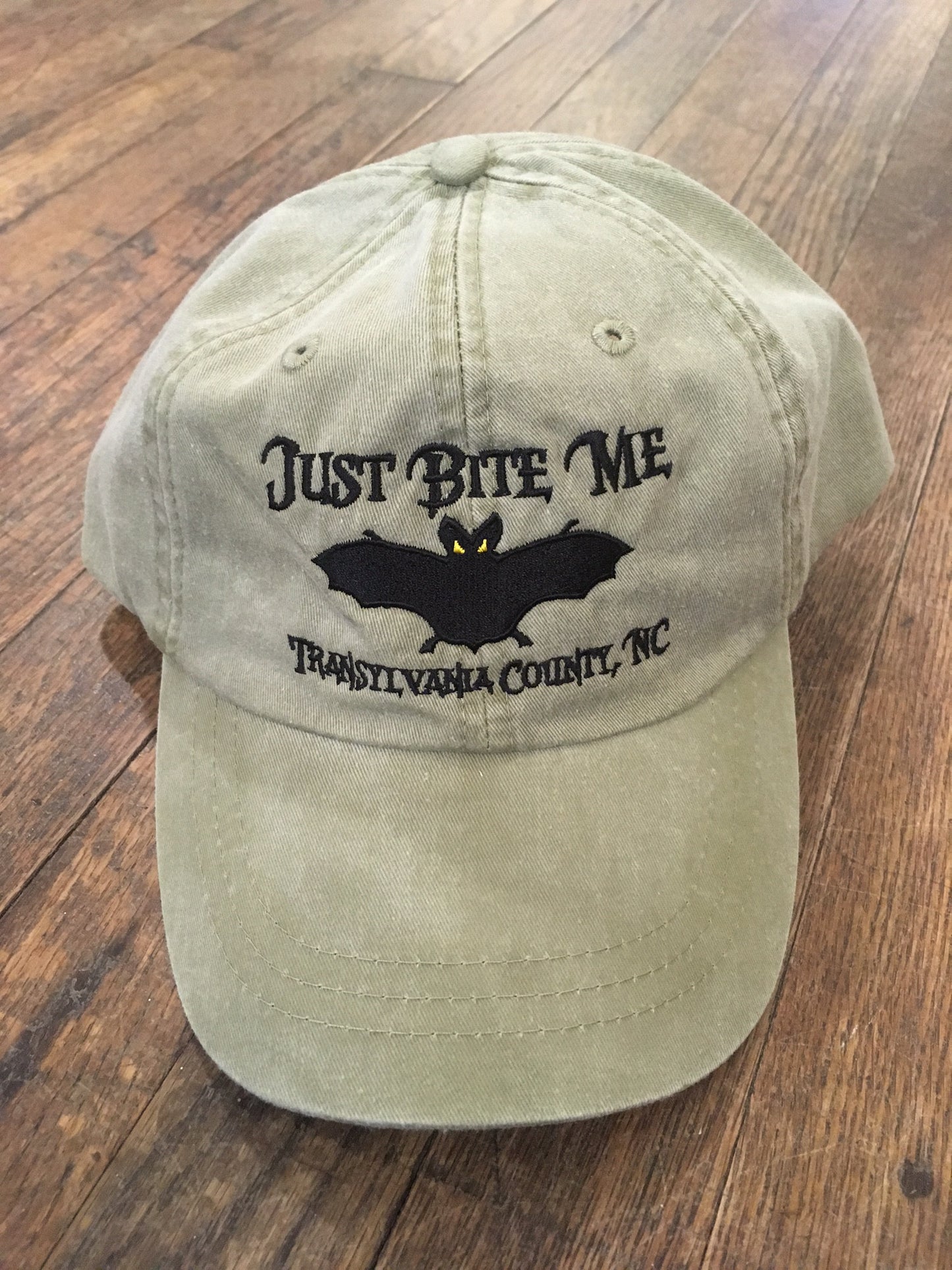A khaki baseball cap with 'Just Bite Me' embroidered on the front and a bat silhouette and Transylvania County, NC.